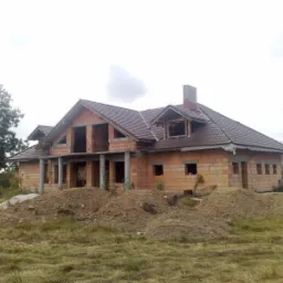Niedokończony, ceglany dom jednorodzinny z brązowym dachem, widoczne kolumny podtrzymujące zadaszenie, hałdy ziemi przed budynkiem, trawa i drzewa w tle.