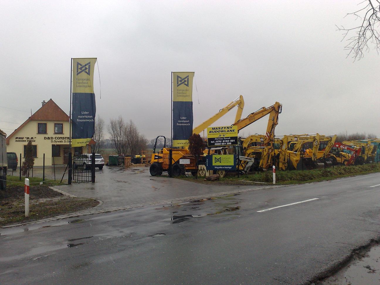 Plac firmy z maszynami budowlanymi Komatsu, widoczne banery reklamowe i budynek biurowy w pochmurny dzień po deszczu.