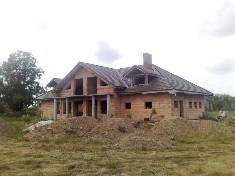 Niedokończony, ceglany dom jednorodzinny z brązowym dachem, widoczne kolumny podtrzymujące zadaszenie, hałdy ziemi przed budynkiem, trawa i drzewa w tle.