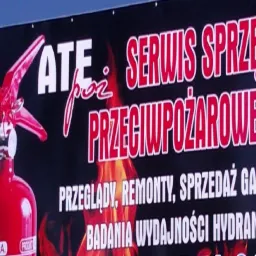 Baner reklamowy z czerwonym gaśnicą, informujący o serwisie sprzętu przeciwpożarowego, przeglądach, remontach, sprzedaży gaśnic i badaniach wydajności hydrantów.