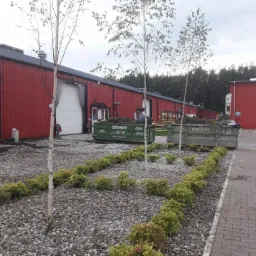 Dwa kontenery wypełnione ziemią i roślinami stojące na żwirowym podłożu przed czerwonymi budynkami przemysłowymi, z młodymi drzewkami i niskimi krzewami, całość w pochmurny dzień.