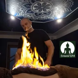 Mężczyzna w maseczce ochronnej wykonuje zabieg z użyciem ognia na plecach klienta leżącego na stole w ciemnym pomieszczeniu z dekoracyjnym sufitem i logo Oriental SPA.