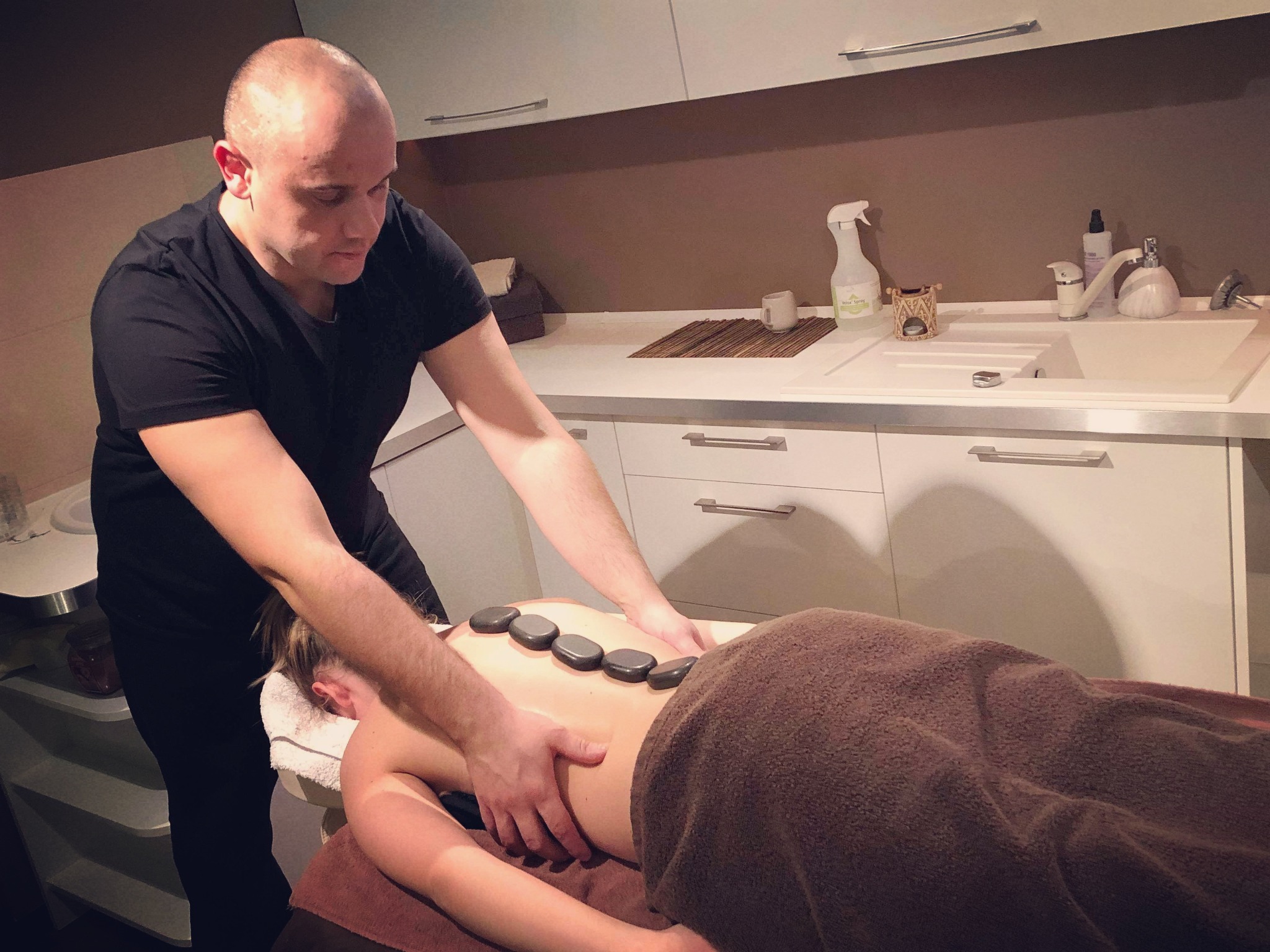 Mężczyzna w czarnej koszulce wykonuje masaż gorącymi kamieniami na plecach kobiety leżącej na ręczniku w gabinecie zabiegowym.