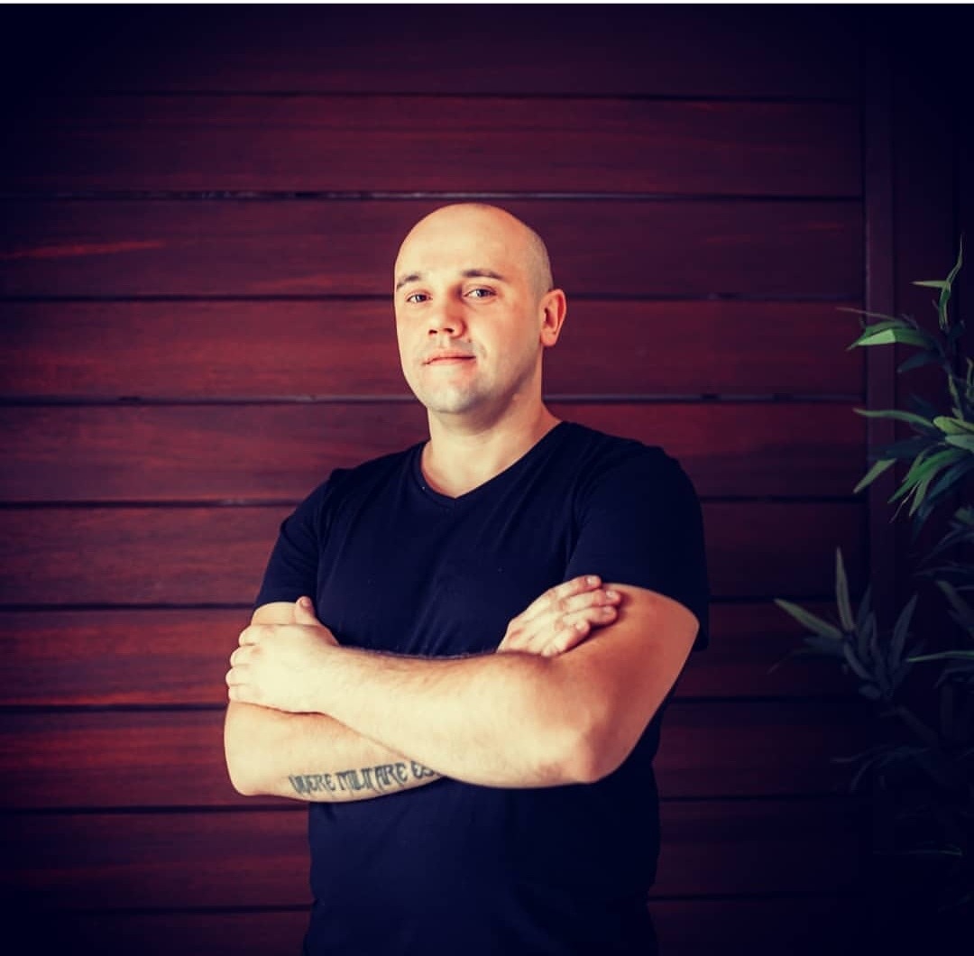 Portret mężczyzny z ogoloną głową, ubranego w czarny t-shirt, z tatuażem na przedramieniu, stojącego na tle ściany z ciemnego drewna i rośliny doniczkowej.