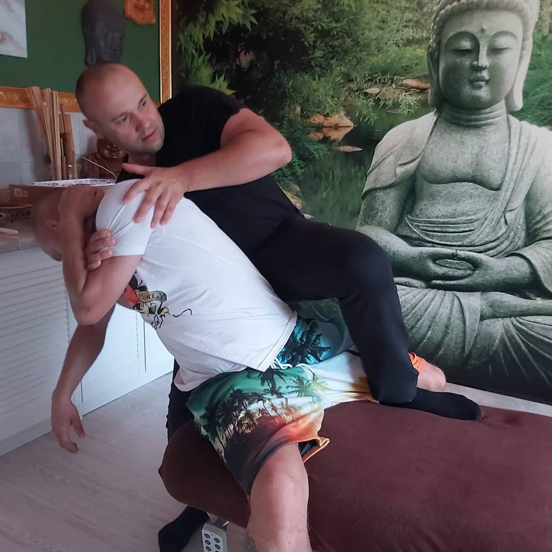 Mężczyzna w czarnej koszulce wykonuje uścisk na pacjencie leżącym na brązowym stole do masażu, w tle tapeta z wizerunkiem Buddy i zieleni.