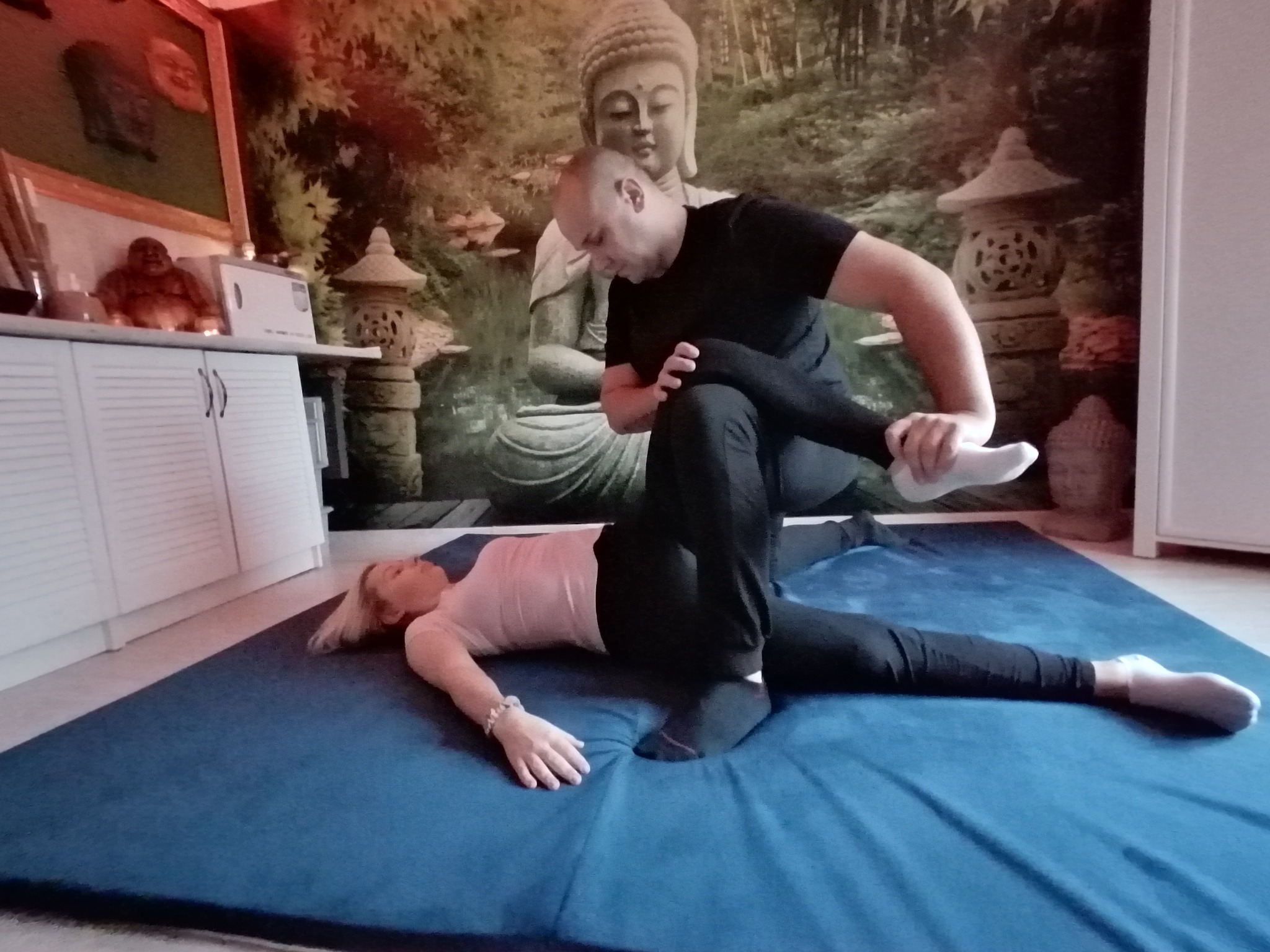 Terapeuta w czarnym stroju wykonuje zabieg na leżącej kobiecie na niebieskiej macie; w tle ściana z motywem Buddy i lampionów.