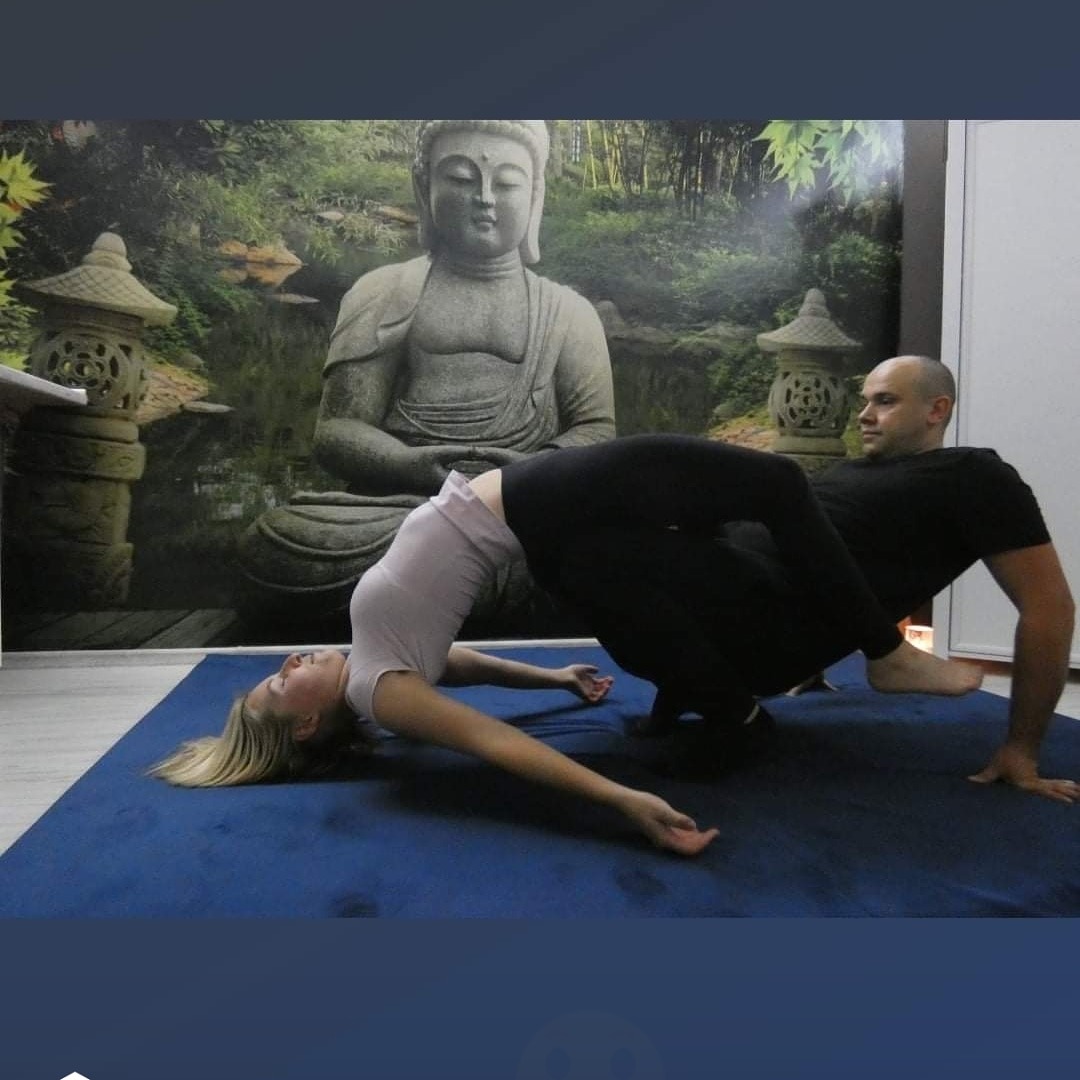 Kobieta w pozycji mostka, wspierana przez mężczyznę, na tle ściany z wizerunkiem Buddy i lampionów.