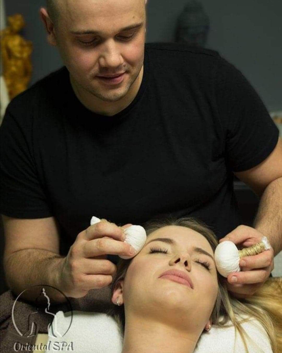 Kobieta leżąca na plecach podczas zabiegu z użyciem białych, ziołowych kompresów trzymanych przez mężczyznę w czarnym T-shircie. Widoczne logo Oriental SPA w lewym dolnym rogu.