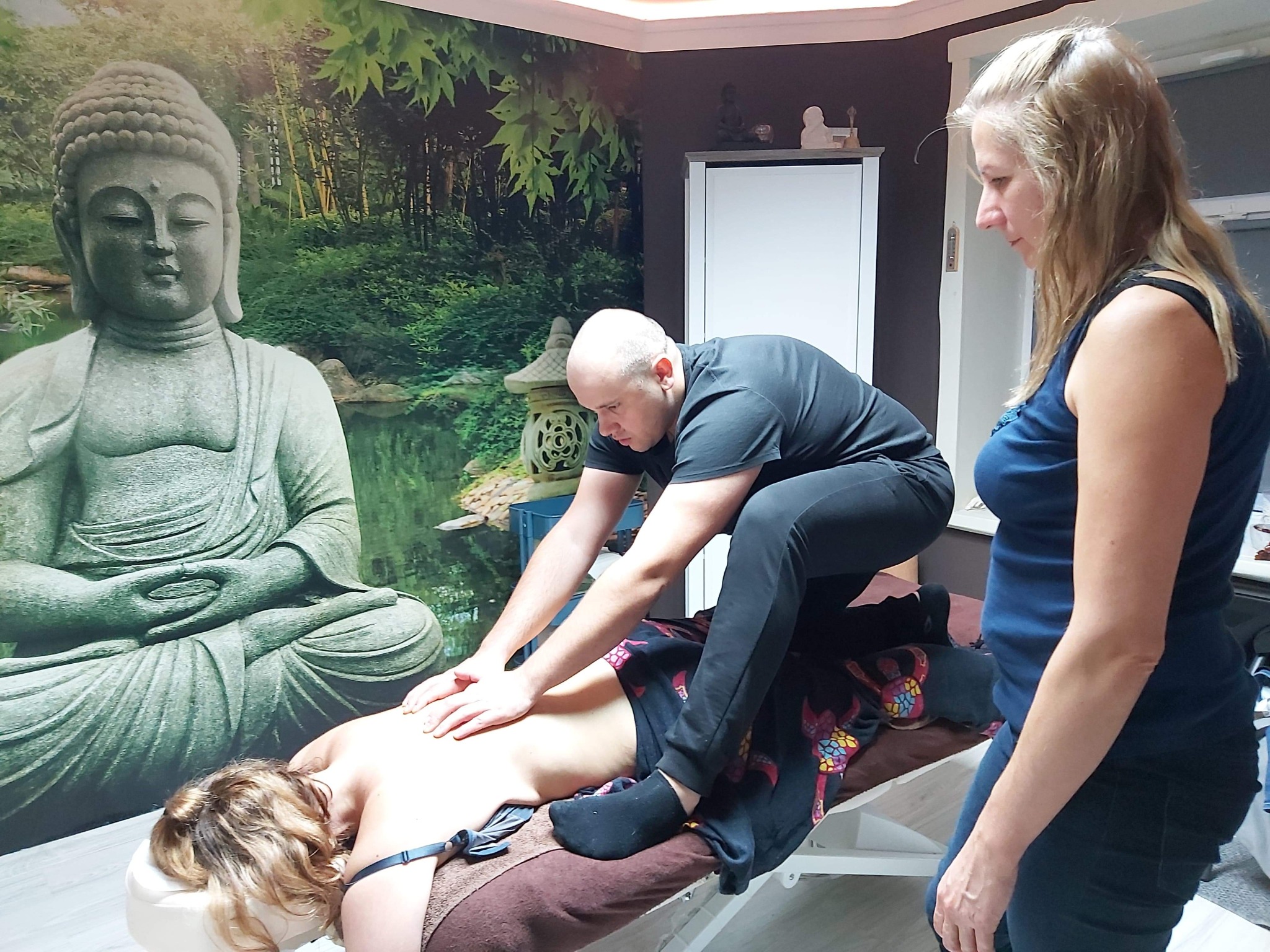 Mężczyzna w czarnym stroju wykonuje masaż pleców kobiecie leżącej na stole do masażu w gabinecie z fototapetą przedstawiającą Buddę i zielony krajobraz; obok stoi kobieta.