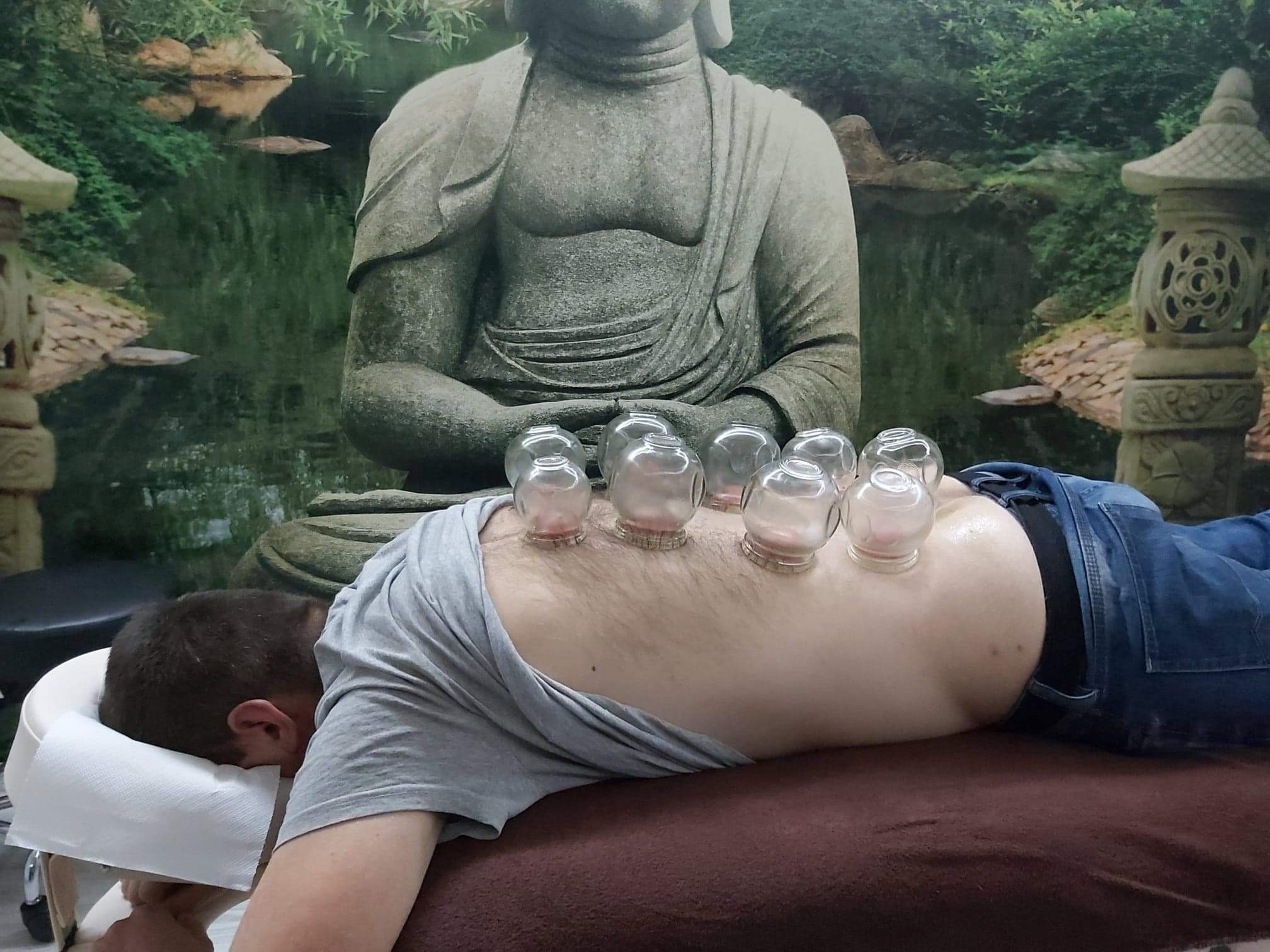 Mężczyzna leżący na brzuchu z przyssawkami na plecach podczas zabiegu bańkoterapii; w tle dekoracyjna tapeta z motywem Buddy, stawu i lampionów.