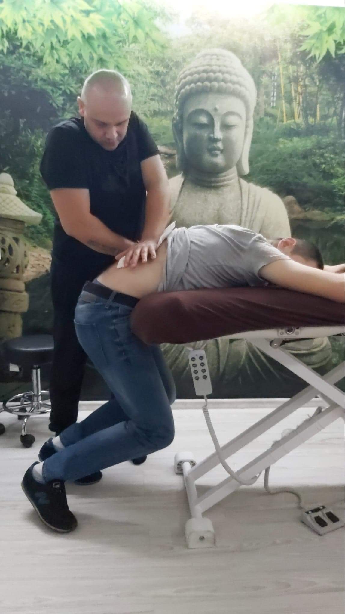 Mężczyzna w czarnej koszulce wykonuje terapię manualną na dolnej części pleców klienta leżącego na specjalistycznym stole z regulacją wysokości, w tle tapeta z motywem Buddy i zieleni.