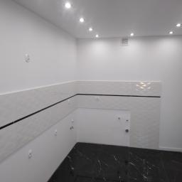 Remoncik - Nowoczesna łazienka po remoncie: białe płytki z geometrycznym wzorem i czarnym paskiem, czarna marmurowa podłoga, oświetlenie punktowe w suficie.