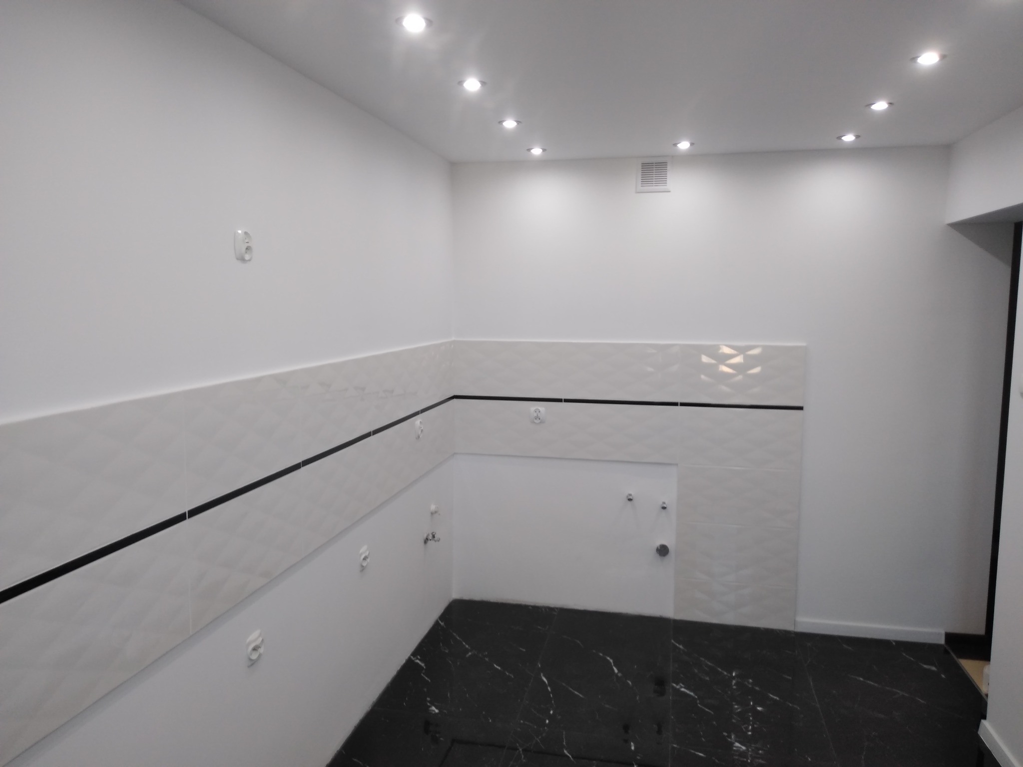 Nowoczesna łazienka po remoncie: białe płytki z geometrycznym wzorem i czarnym paskiem, czarna marmurowa podłoga, oświetlenie punktowe w suficie.