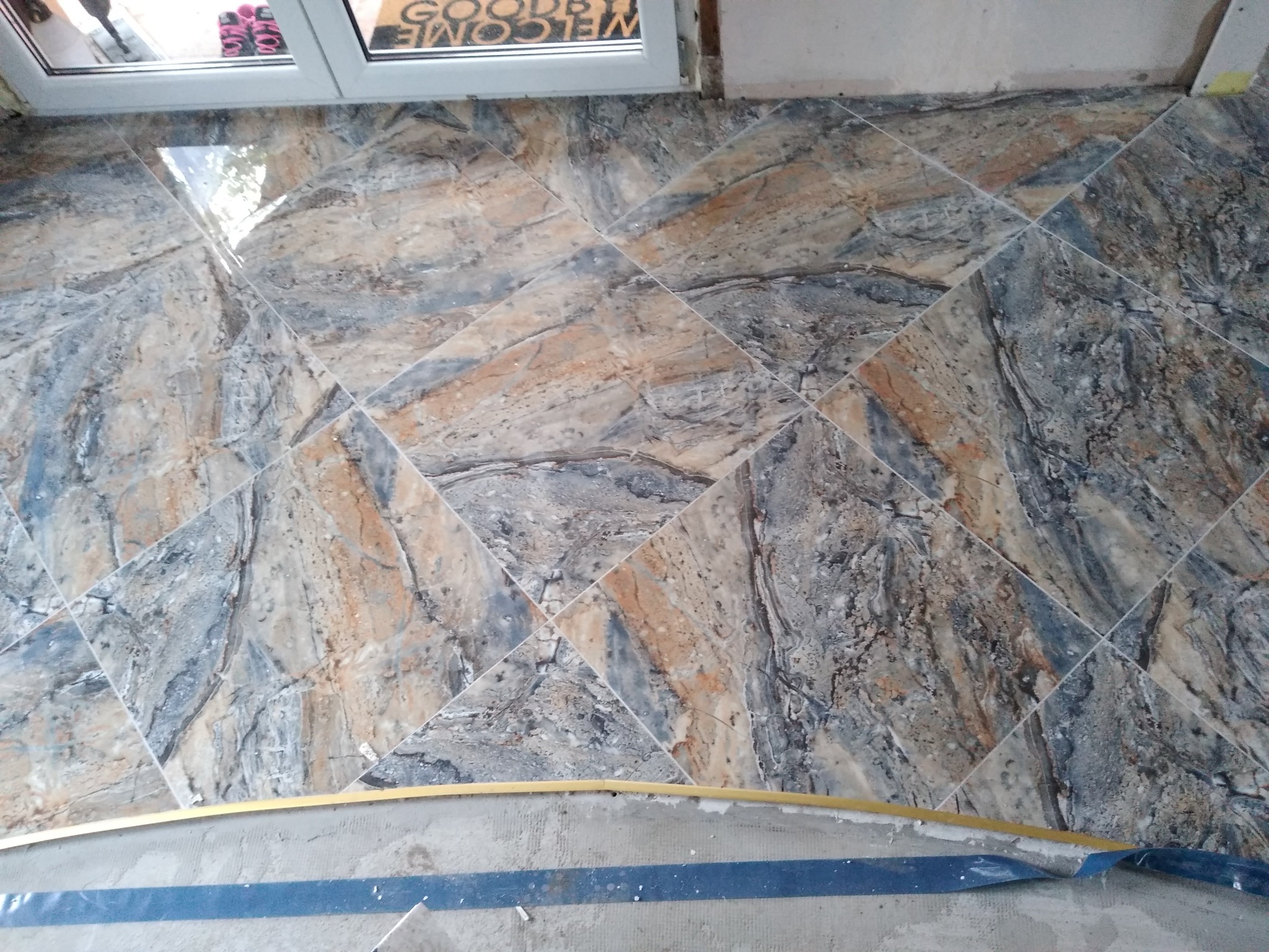 Ułożona pod kątem podłoga z płytek imitujących marmur w odcieniach szarości, beżu i brązu, widoczna fuga, okno w tle, zabezpieczona taśmą krawędź podłogi.