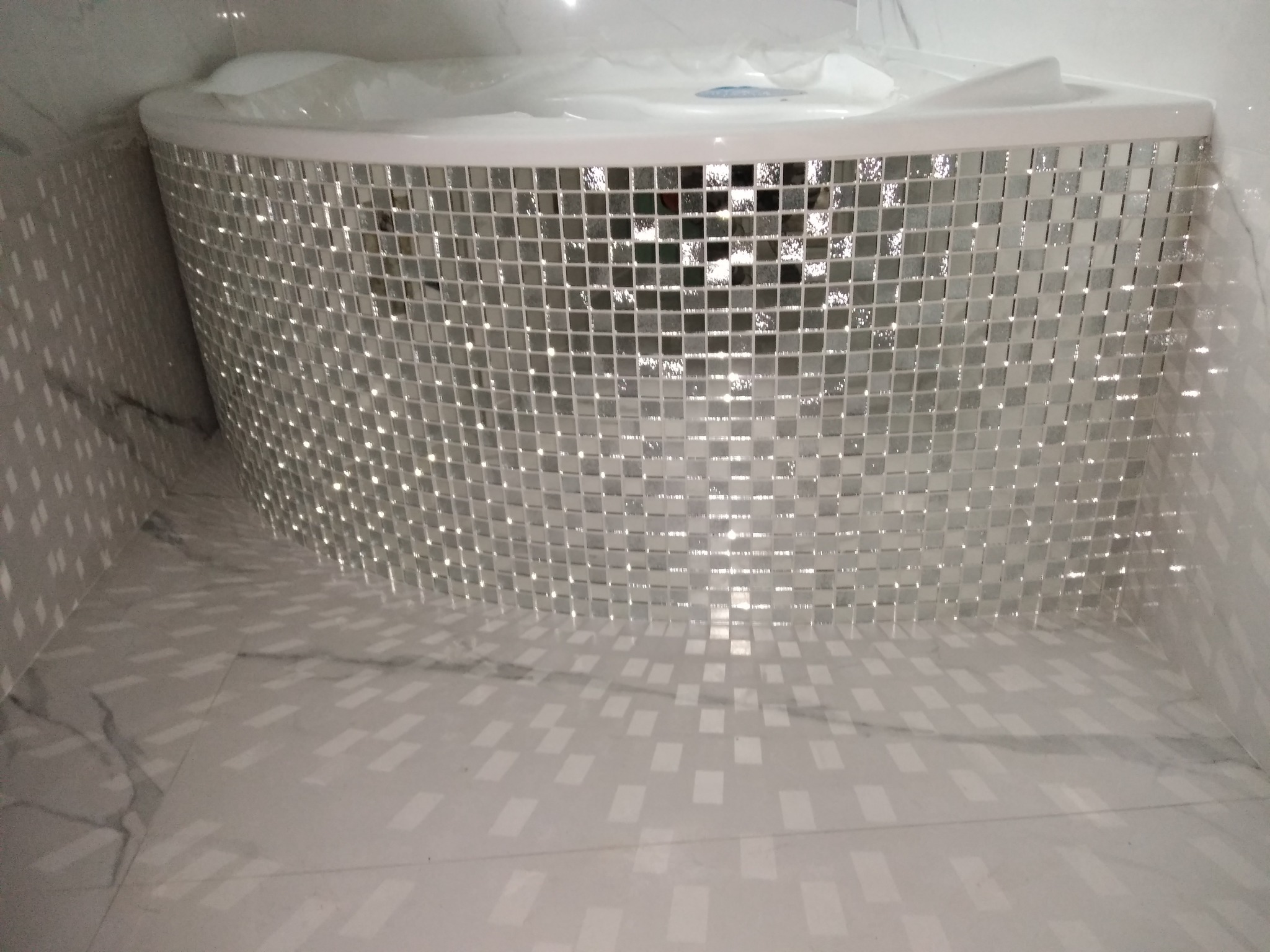 Błyszcząca wanna narożna obłożona mozaiką z małych, srebrnych płytek, odbijających światło na marmurowej podłodze w łazience.