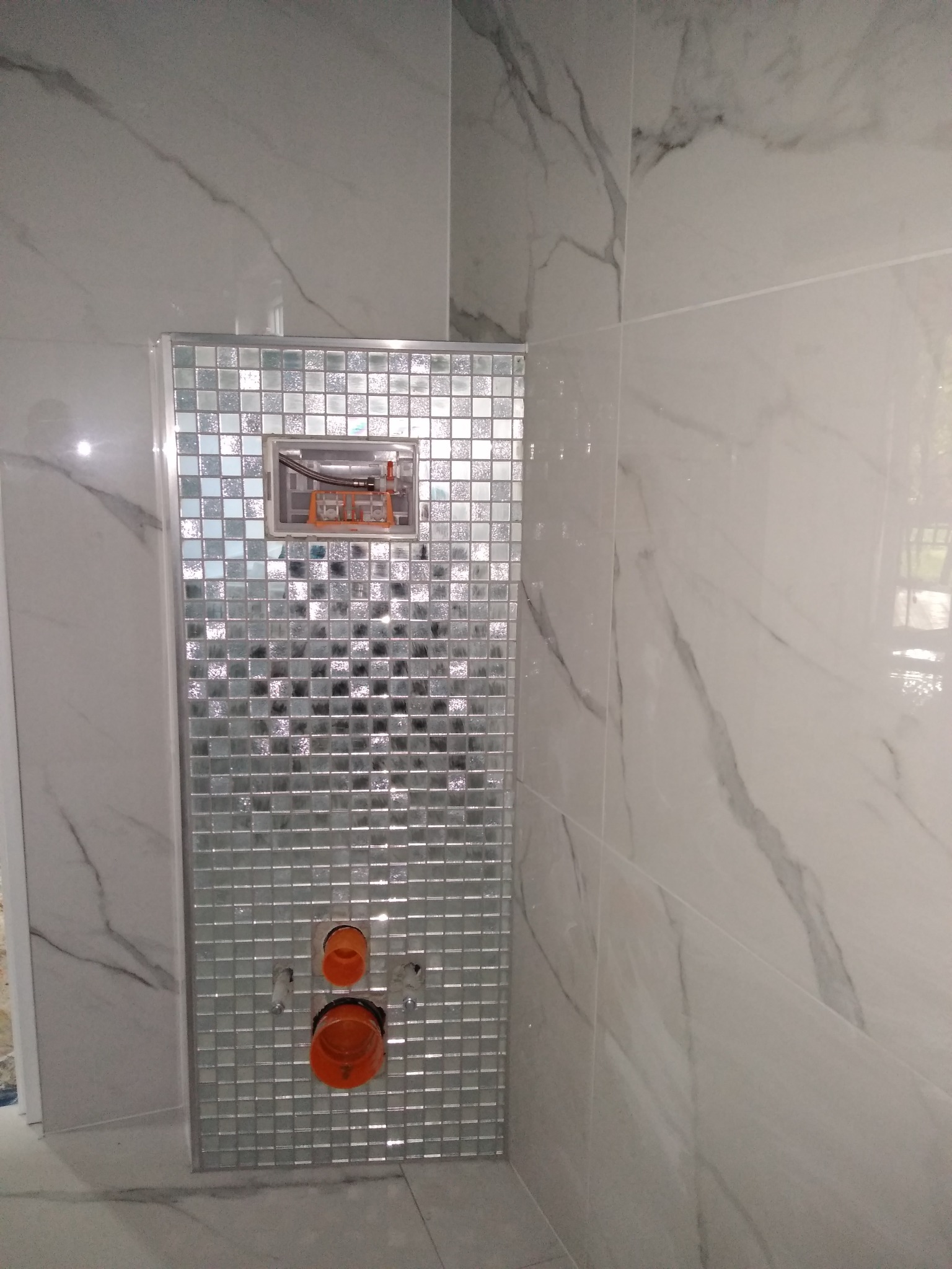 Błyszcząca mozaika z drobnymi kwadratami na zabudowie stelaża podtynkowego WC, widoczne pomarańczowe rury i białe marmurowe płytki na ścianie.