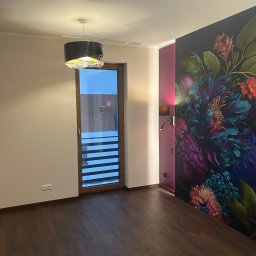 UBERR Renata Rakowska - Pokój po remoncie z ciemną podłogą, ścianą z motywem kwiatowym i lampą wiszącą w folii. Widoczne gniazdka elektryczne i drzwi balkonowe z żaluzjami.