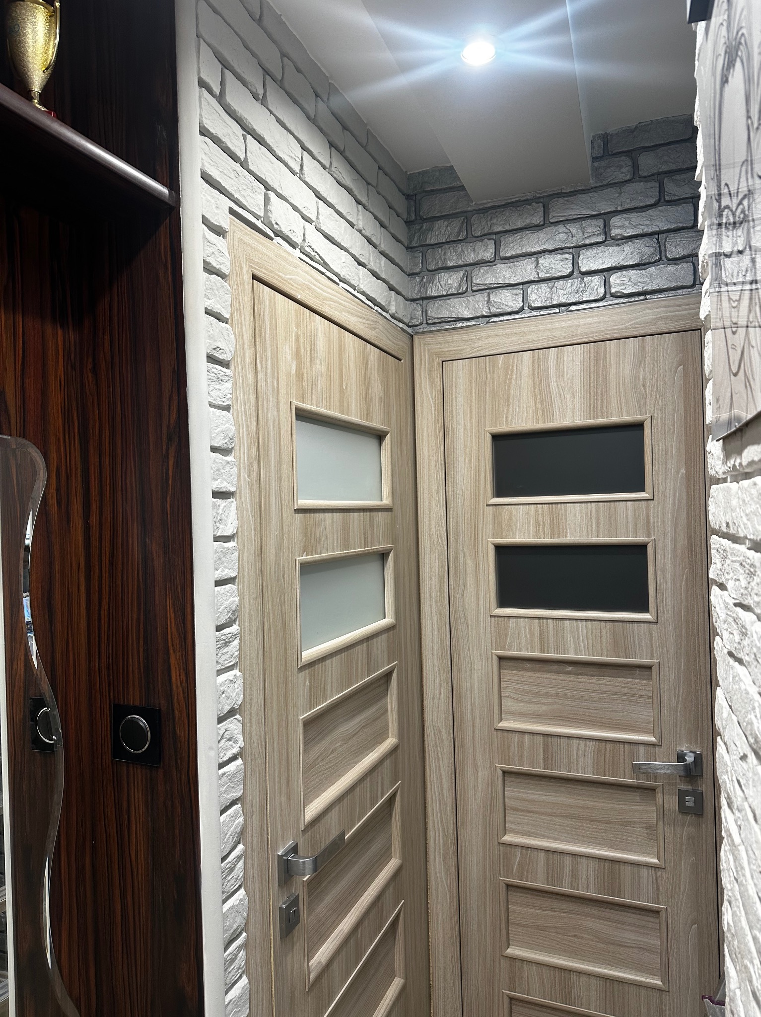 Dwa różne modele drzwi wewnętrznych z przeszkleniami, zamontowane w pomieszczeniu z dekoracyjną ścianą z białej cegły. Widoczne klamki i fragment szafy z ciemnego drewna.