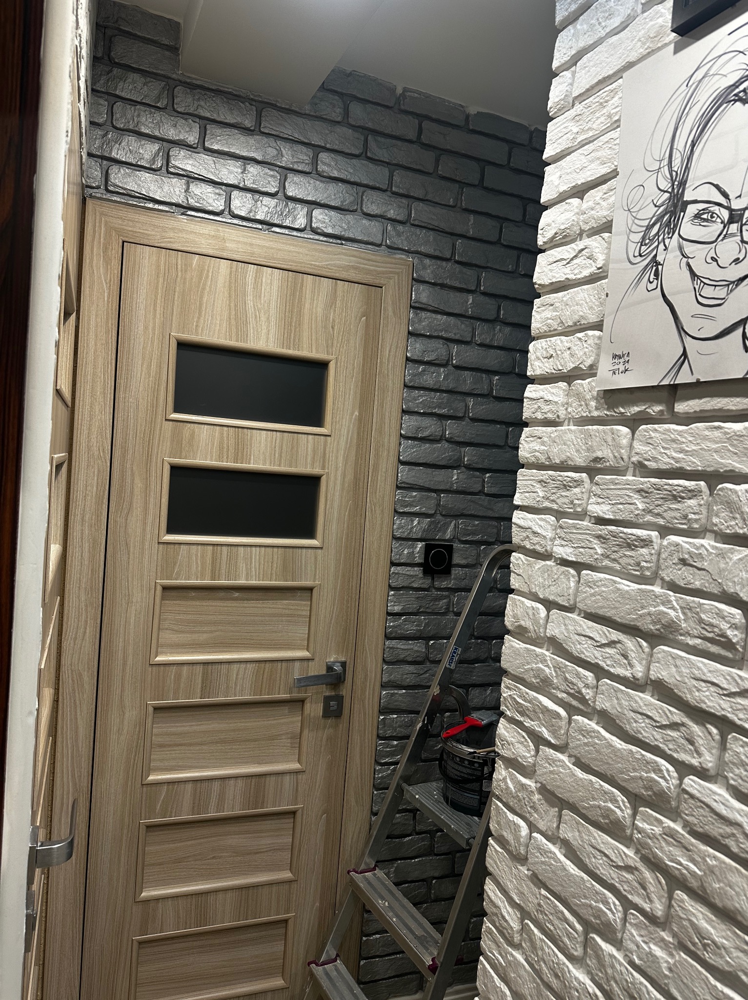 Wnętrze po remoncie: drewniane drzwi z szybkami, ściana z szarej cegły i biała ściana z cegły z rysunkiem. Widoczna drabina i wiadro z farbą.