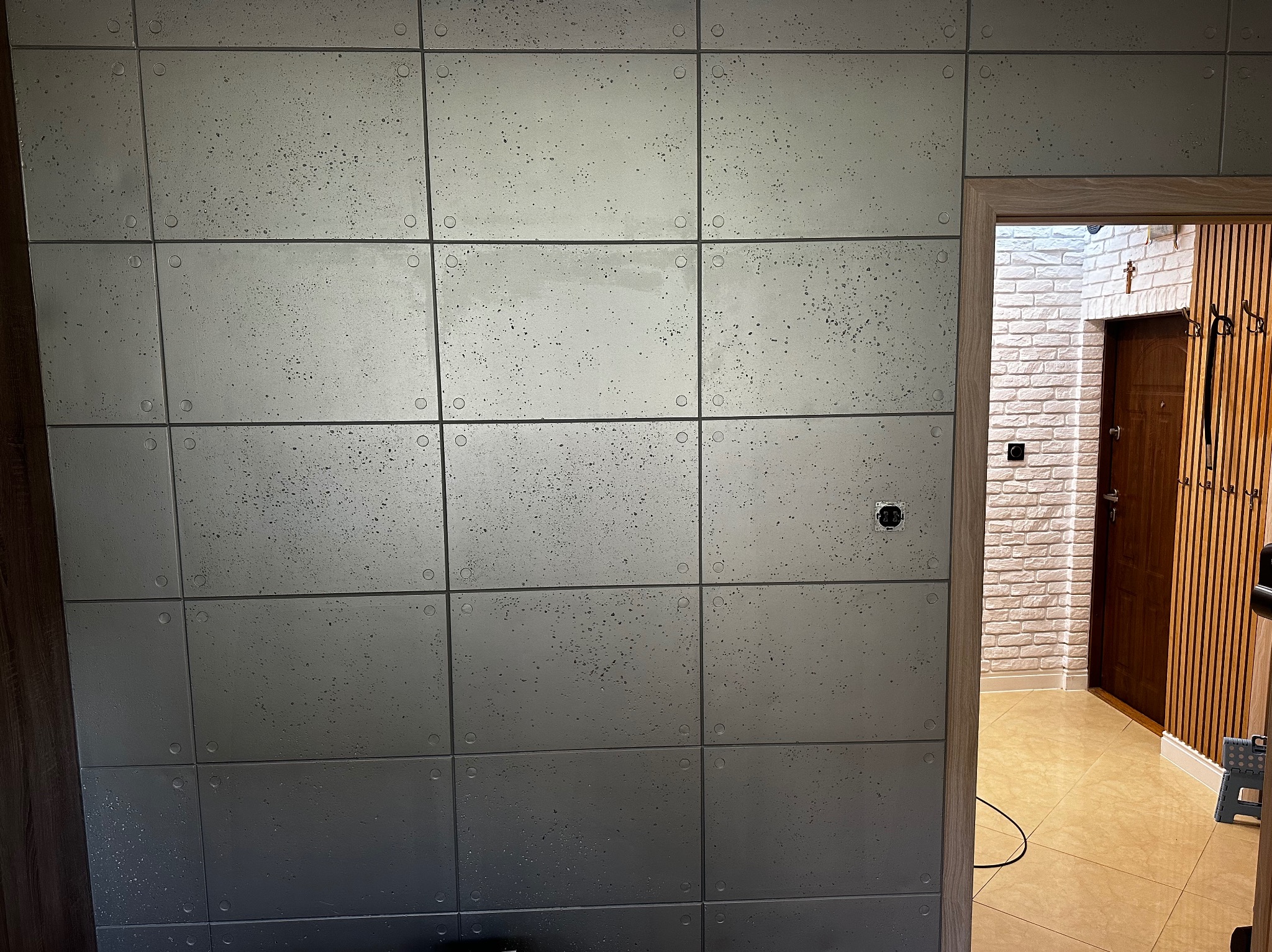 Ściana wykończona panelami imitującymi beton architektoniczny, widoczny fragment instalacji elektrycznej i wnętrze z cegłą oraz drewnianymi lamelami w tle.