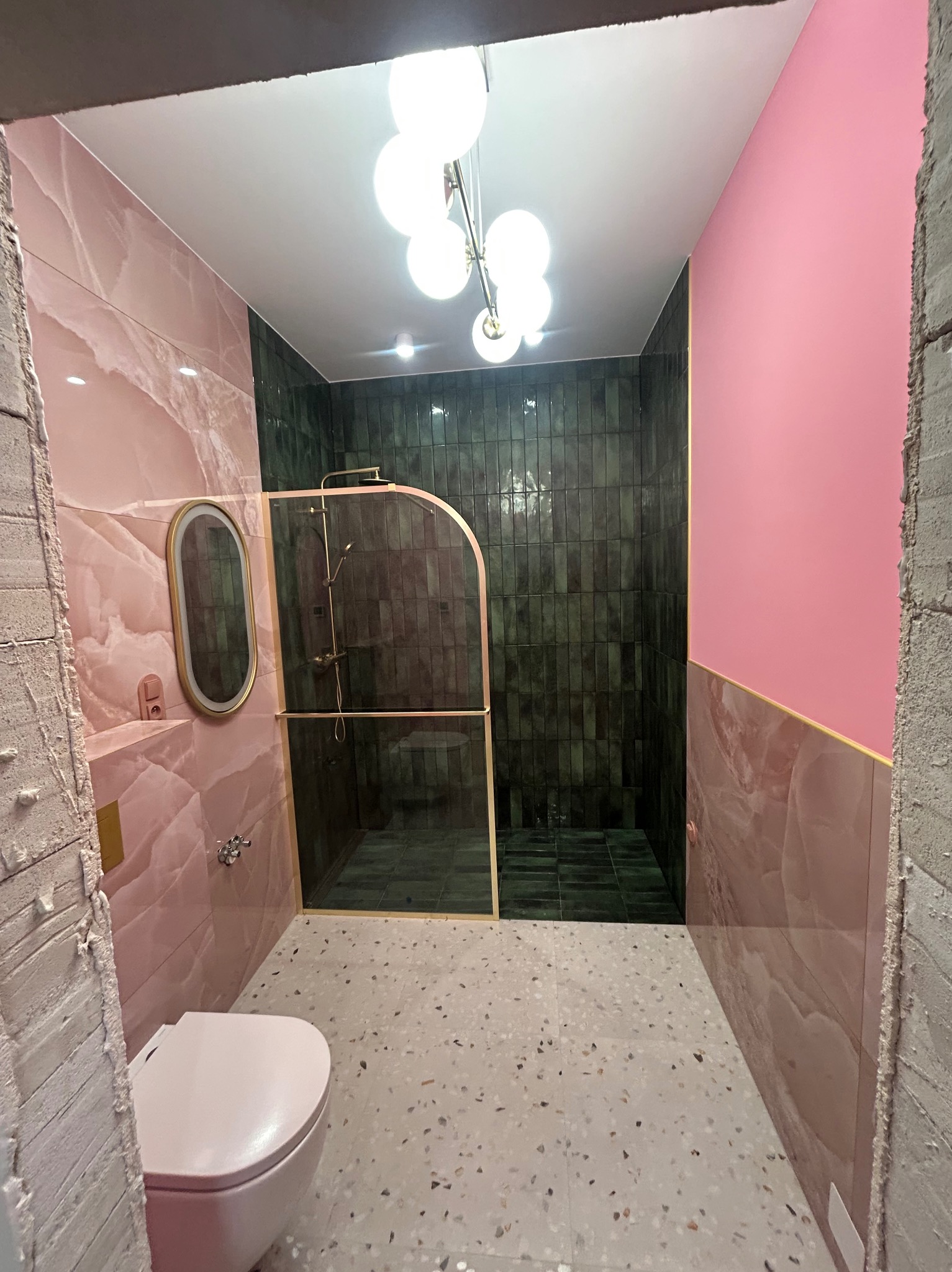 Nowoczesna łazienka w trakcie wykończenia: różowe marmurowe płytki, zielona kabina prysznicowa, terazzo na podłodze, surowy beton przy wejściu. Nietypowe połączenie kolorów.