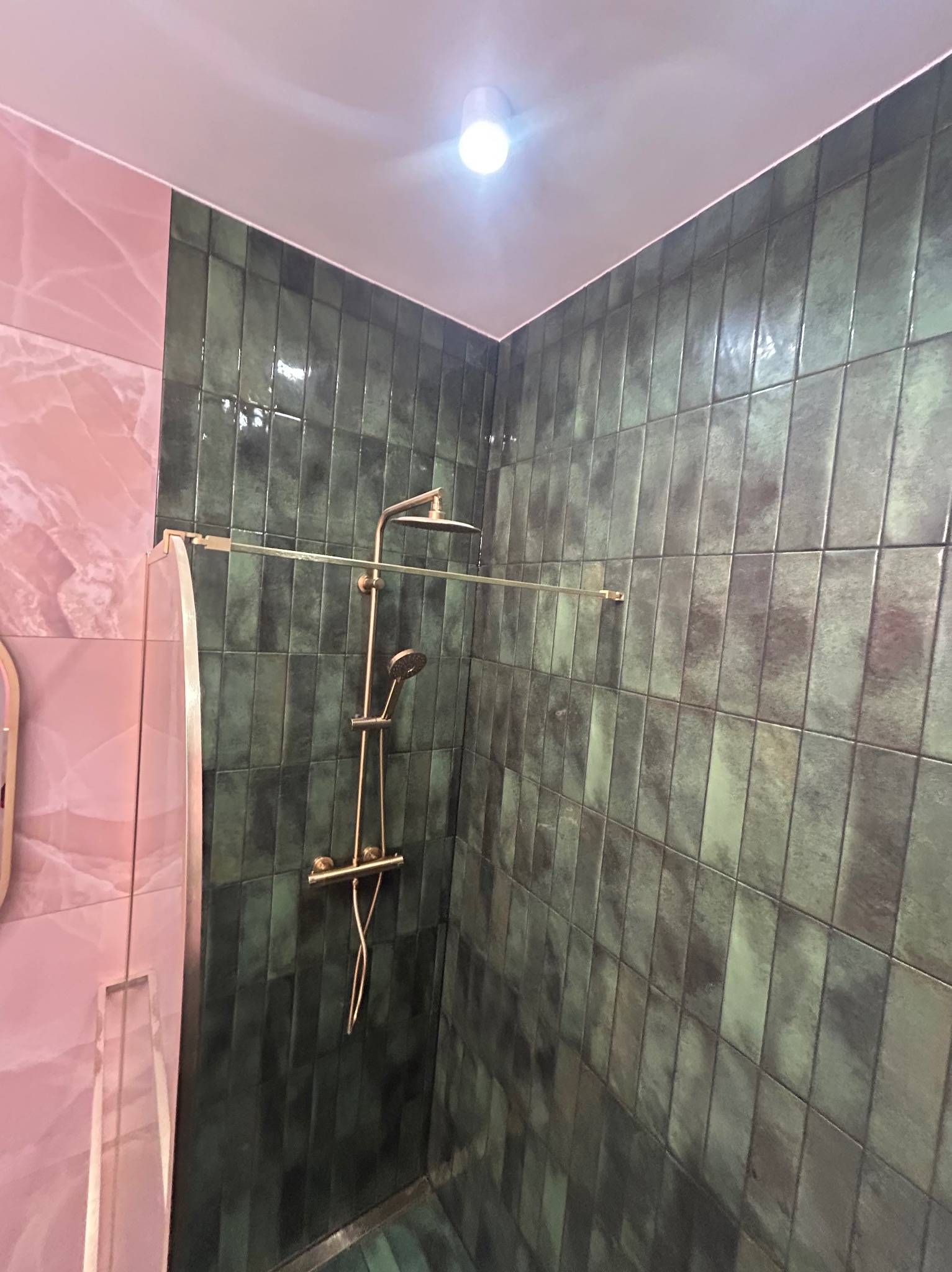 Nowoczesna kabina prysznicowa z zielonymi, błyszczącymi płytkami ułożonymi pionowo, złotą armaturą i różową ścianą z efektem marmuru. Minimalistyczny design.