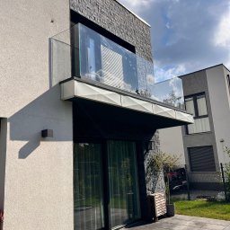 BINGLASS NATALIA RACZYŃSKA - Nowoczesny balkon z szklaną balustradą, widok z dołu, podkreślający minimalistyczny design i użycie szkła. Eleganckie wykończenie budynku z elementami kamiennymi.