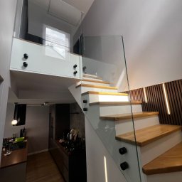 BINGLASS NATALIA RACZYŃSKA - Nowoczesne schody z drewnianymi stopniami i szklaną balustradą, mocowaną czarnymi uchwytami. W tle widoczna kuchnia i dekoracyjna ściana z oświetleniem.