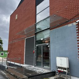 BINGLASS NATALIA RACZYŃSKA - Szklany daszek nad wejściem do budynku z czerwonej cegły. Widoczne mocowania i szklana tafla. Na pierwszym planie skrzynka elektryczna i mokre schody.