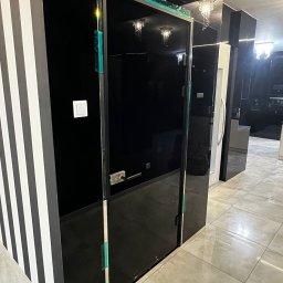 BINGLASS NATALIA RACZYŃSKA - Nowoczesne, czarne drzwi szklane z metalowym uchwytem, w minimalistycznym wnętrzu z elementami w paski i połyskującą podłogą. Widoczne zabezpieczenia na krawędziach.