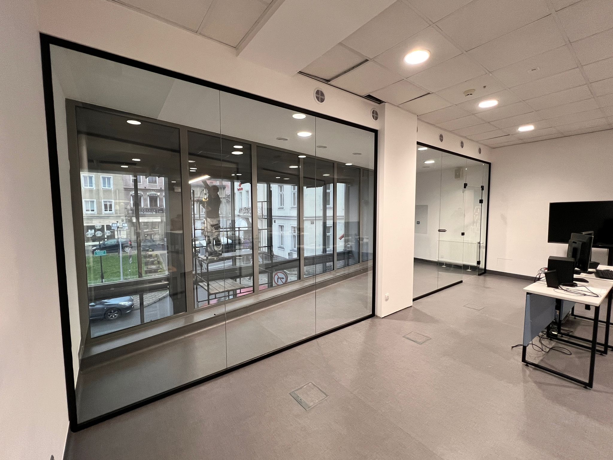 Nowoczesne, przeszklone biuro z widokiem na miasto. Szklane ściany działowe z czarnymi ramami oddzielają przestrzeń. Widoczne biurka i komputery.