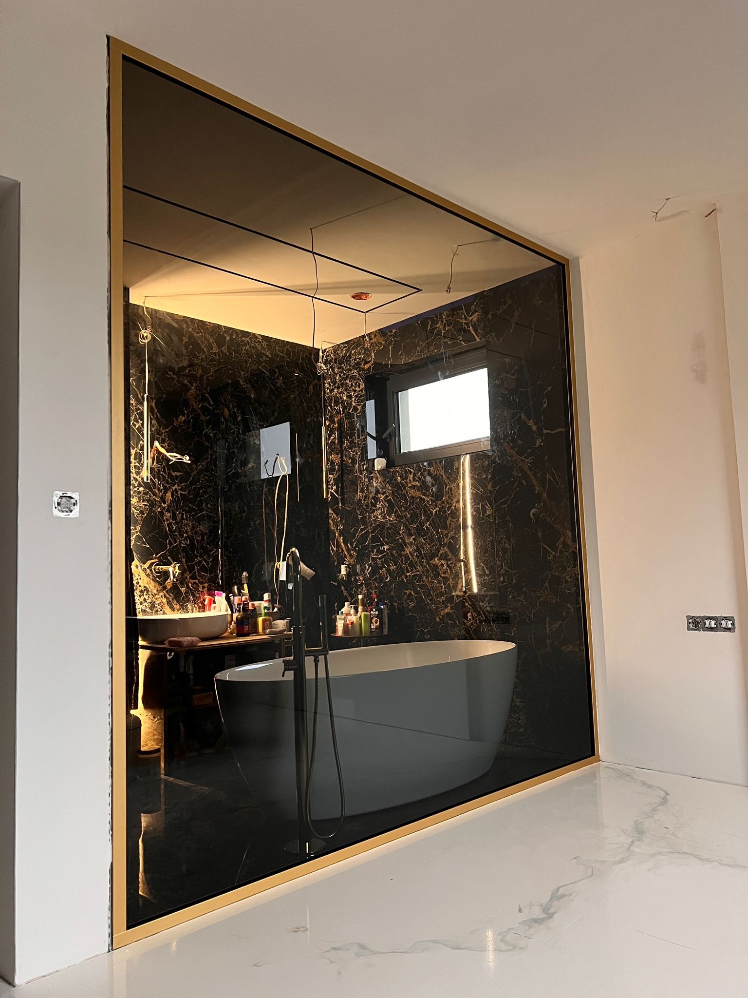 Duże lustro w złotej ramie, odbijające luksusową łazienkę z wanną i marmurowymi ścianami. Widoczne detale wykończenia i oświetlenie.