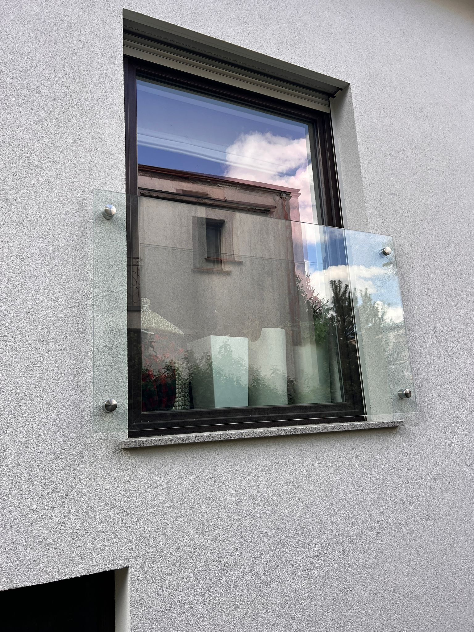 Szklana osłona okienna zamontowana na elewacji budynku, mocowana za pomocą metalowych śrub. W szybie odbija się budynek i niebo.