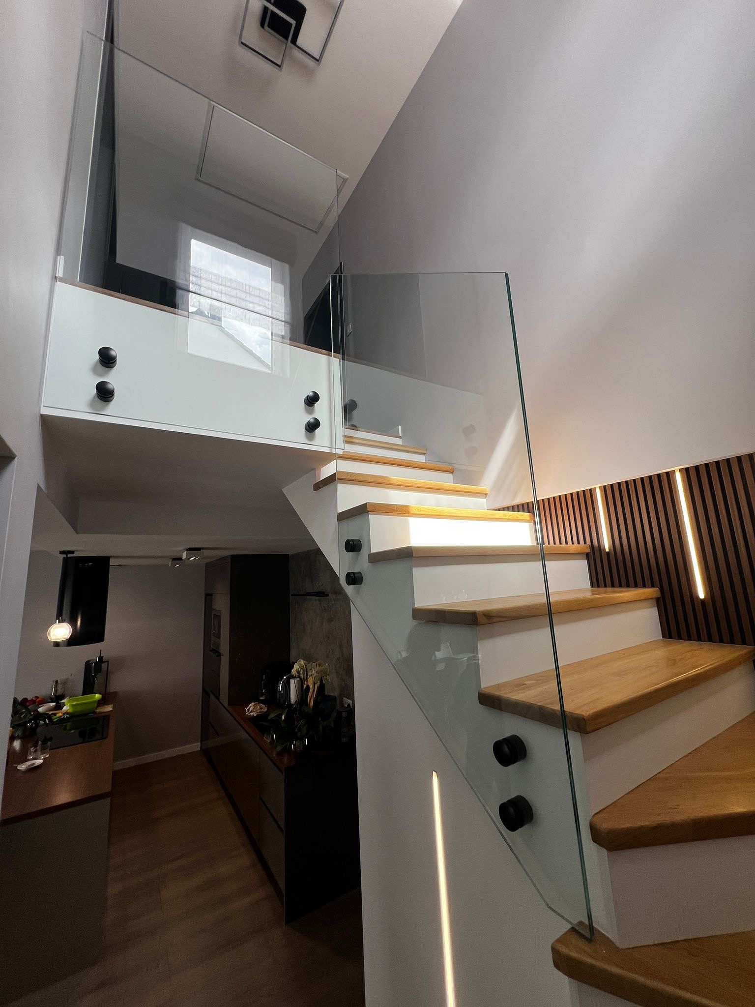 Nowoczesne schody z drewnianymi stopniami i szklaną balustradą, mocowaną czarnymi uchwytami. W tle widoczna kuchnia i dekoracyjna ściana z oświetleniem.