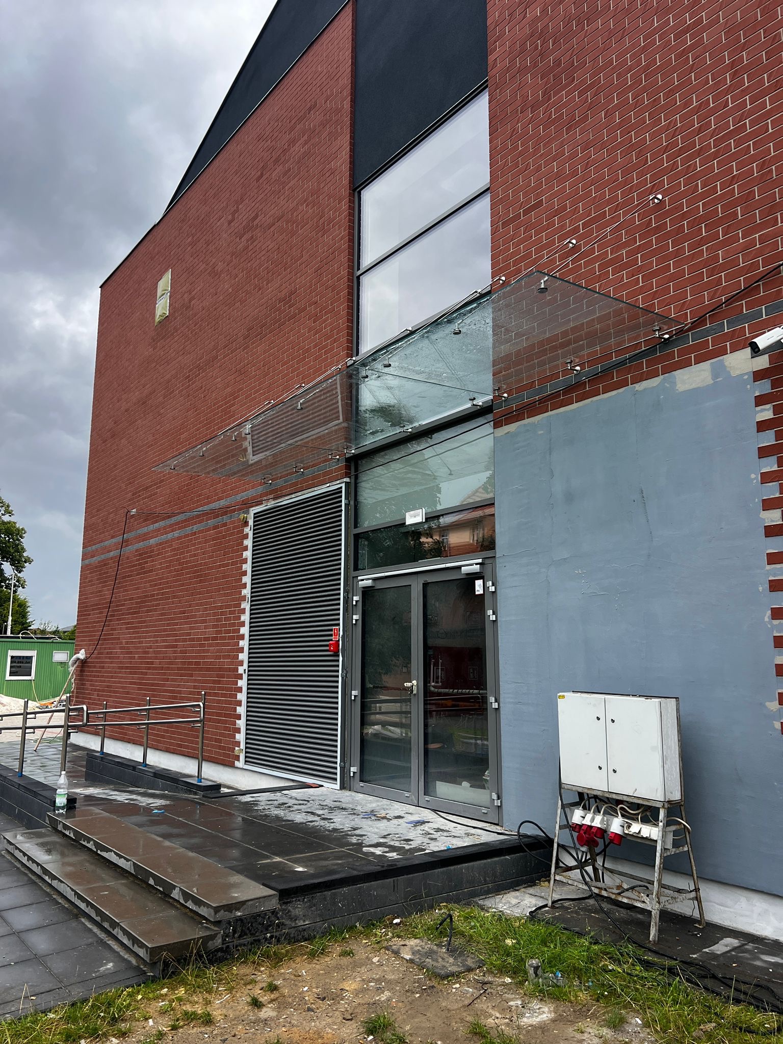 Szklany daszek nad wejściem do budynku z czerwonej cegły. Widoczne mocowania i szklana tafla. Na pierwszym planie skrzynka elektryczna i mokre schody.