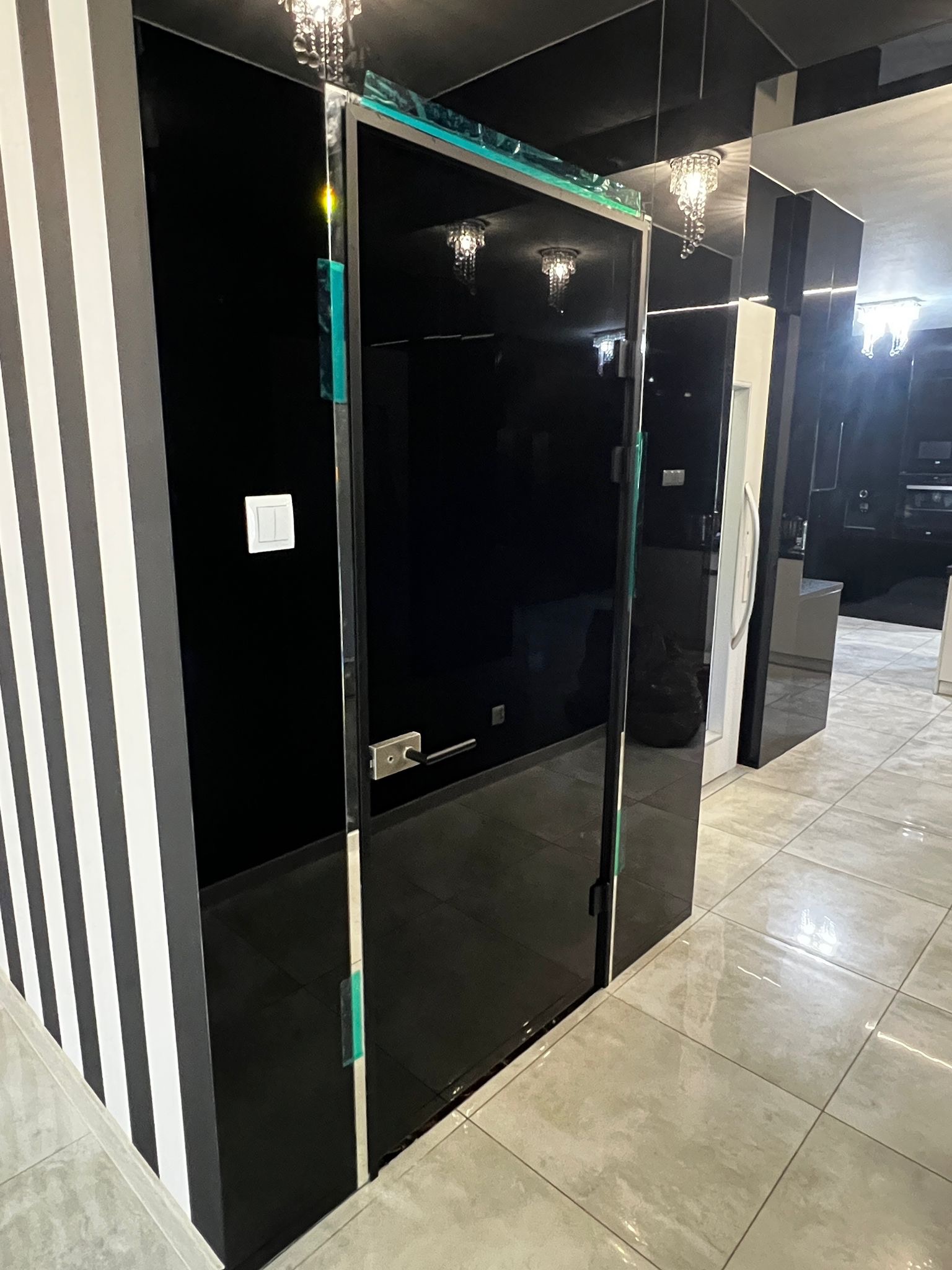 Nowoczesne, czarne drzwi szklane z metalowym uchwytem, w minimalistycznym wnętrzu z elementami w paski i połyskującą podłogą. Widoczne zabezpieczenia na krawędziach.