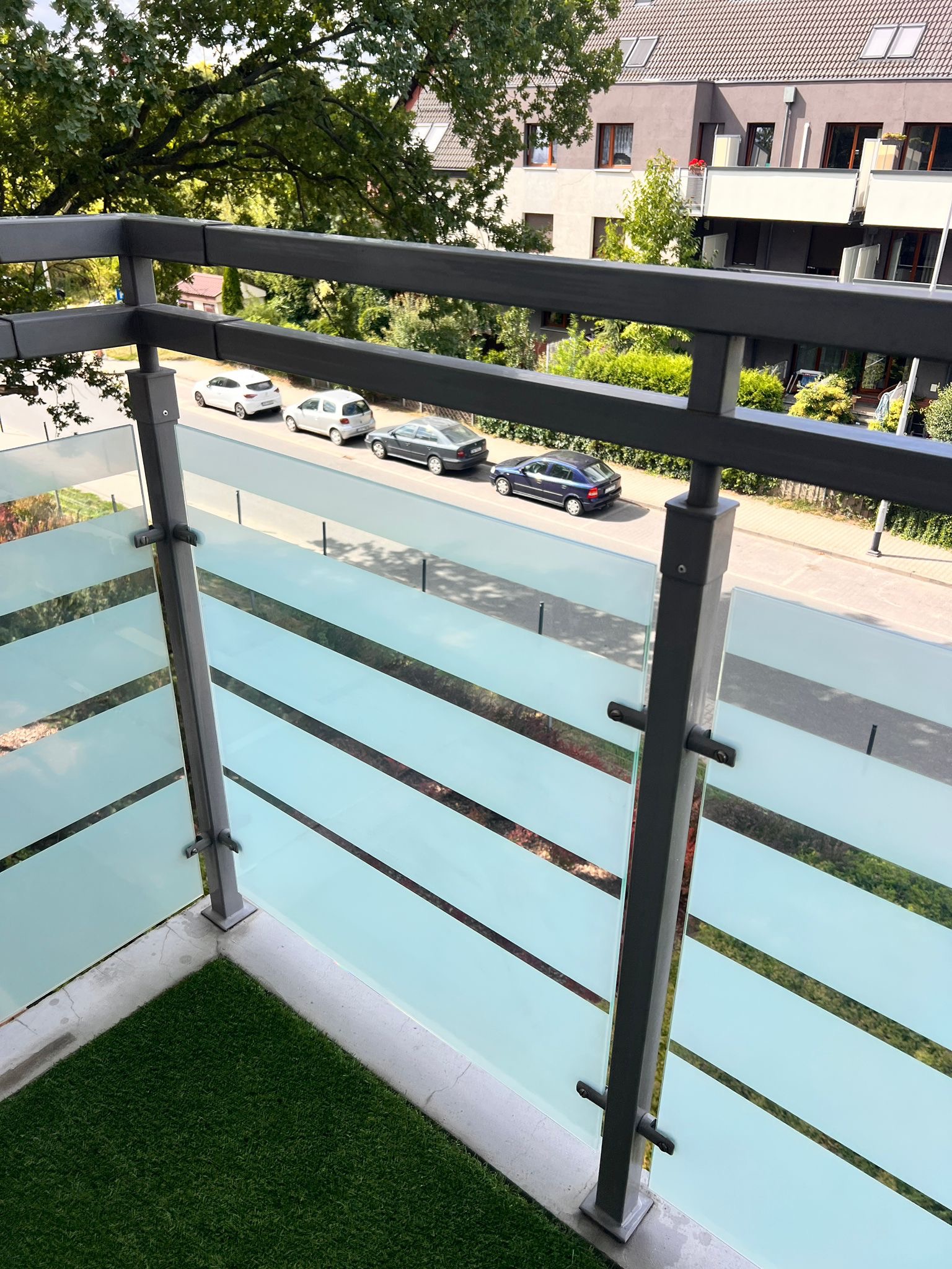 Nowoczesna balustrada balkonowa z matowego szkła i metalową ramą, widok z góry na ulicę z zaparkowanymi samochodami, sztuczna trawa na balkonie.