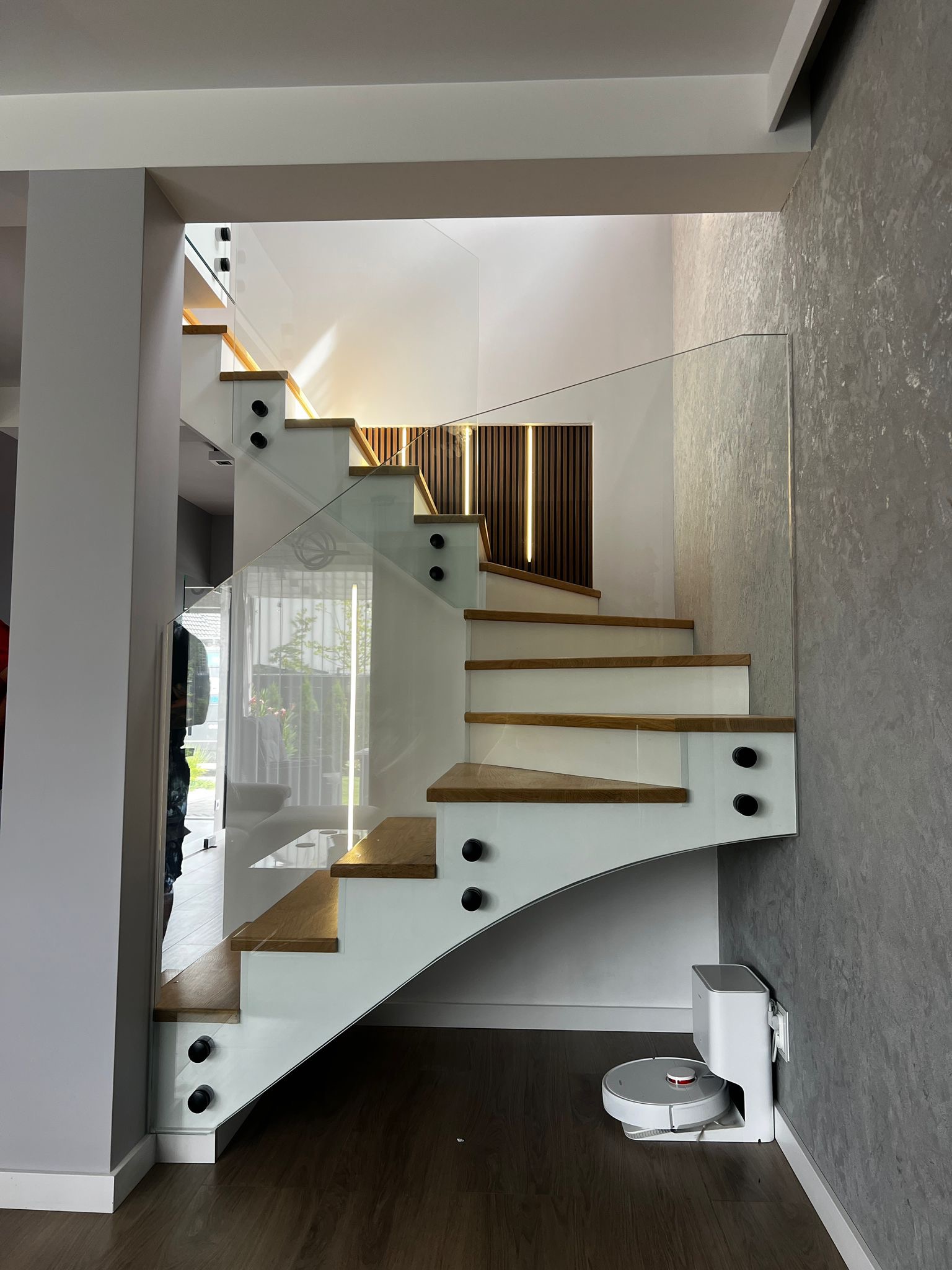 Nowoczesne schody z drewnianymi stopniami i szklaną balustradą, mocowane czarnymi elementami. W tle dekoracyjna ściana z drewnianymi panelami i oświetleniem.