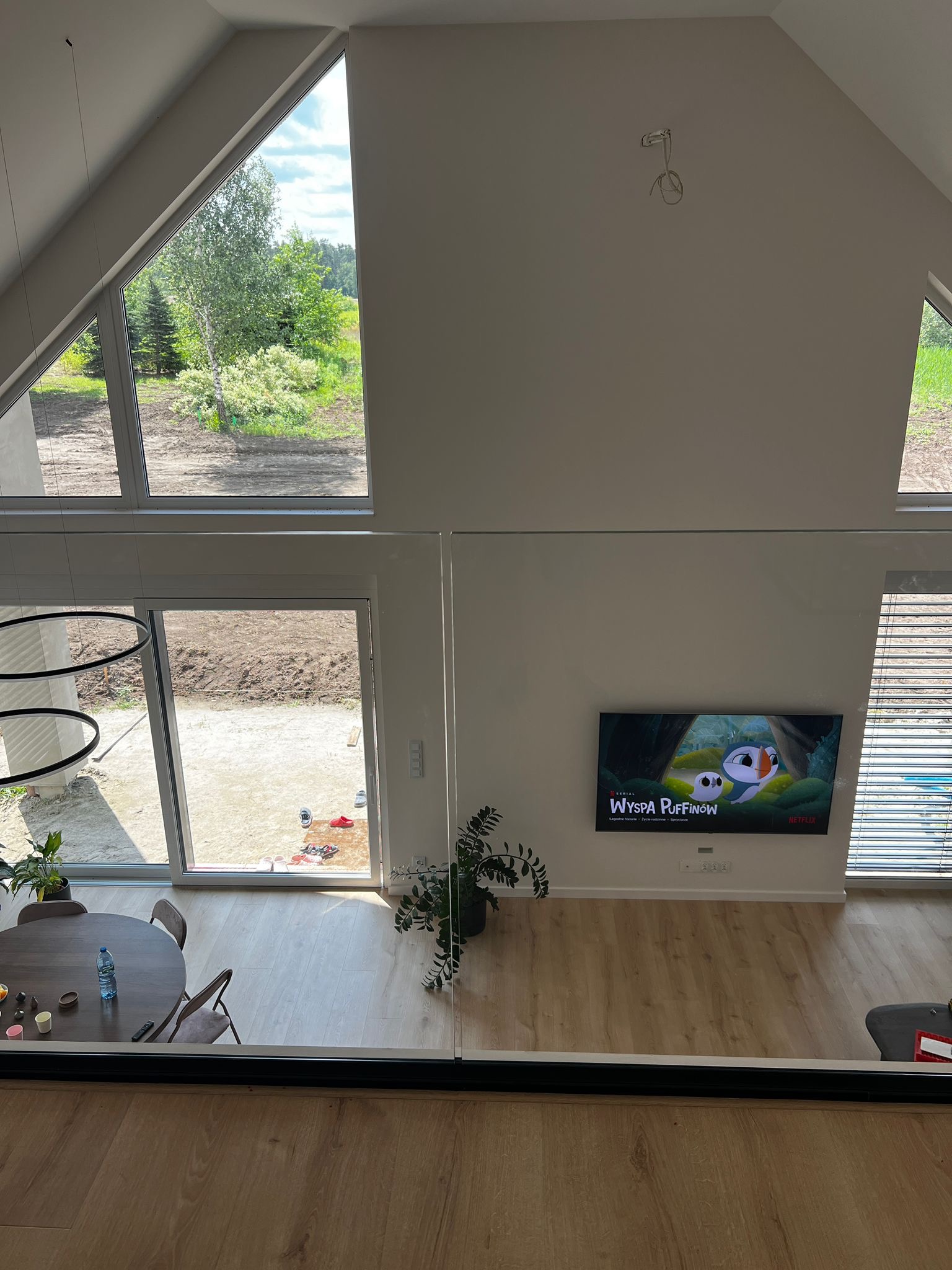 Nowoczesne wnętrze z dużymi oknami i szklaną barierką. Widok z góry na salon z drewnianą podłogą, okrągłym stołem i telewizorem. Duże przeszklenia wpuszczają dużo światła.