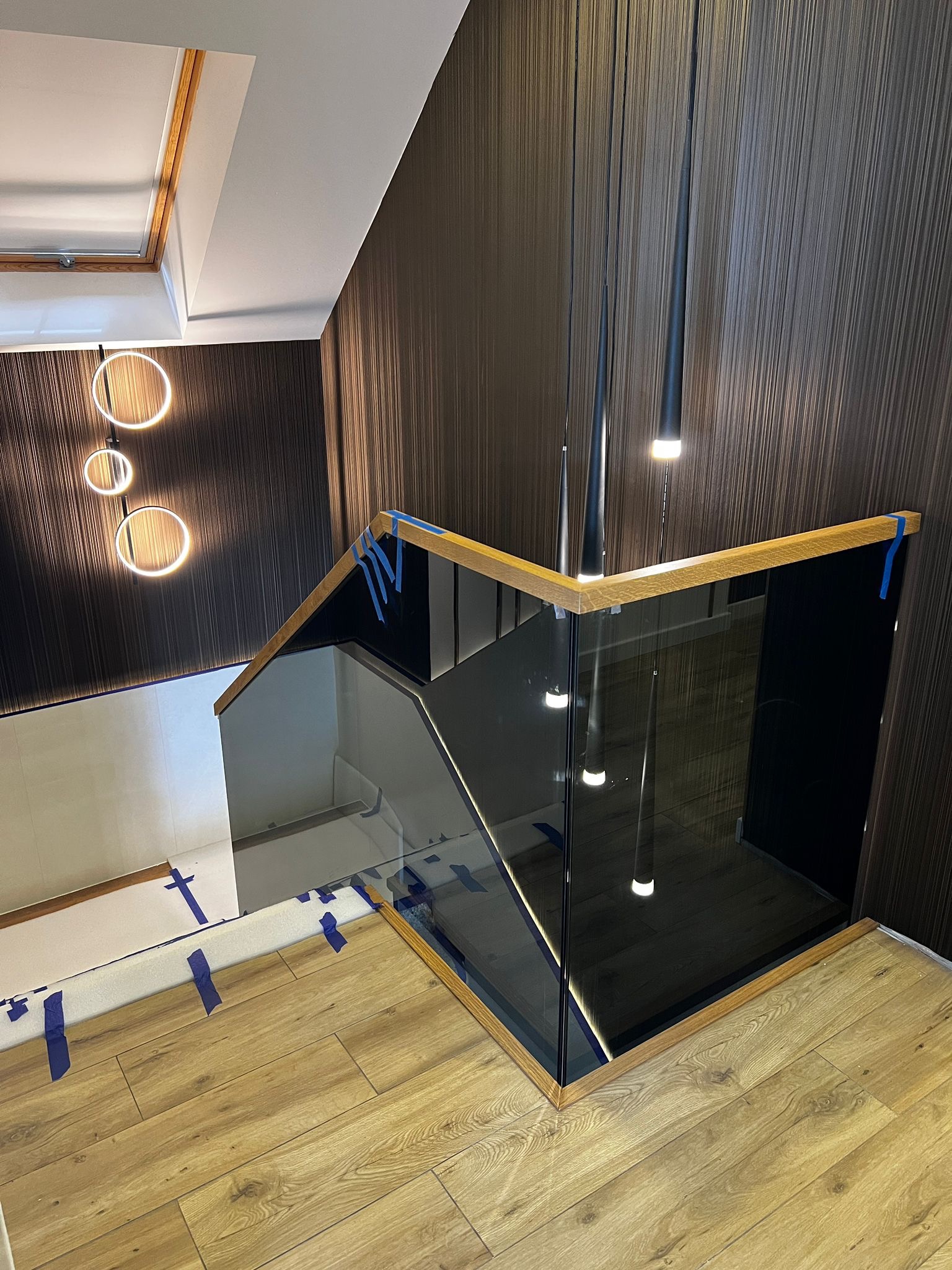 Nowoczesna szklana balustrada schodowa z drewnianymi poręczami, zabezpieczona taśmą malarską. Wiszące lampy i drewniana podłoga uzupełniają elegancki wystrój wnętrza.