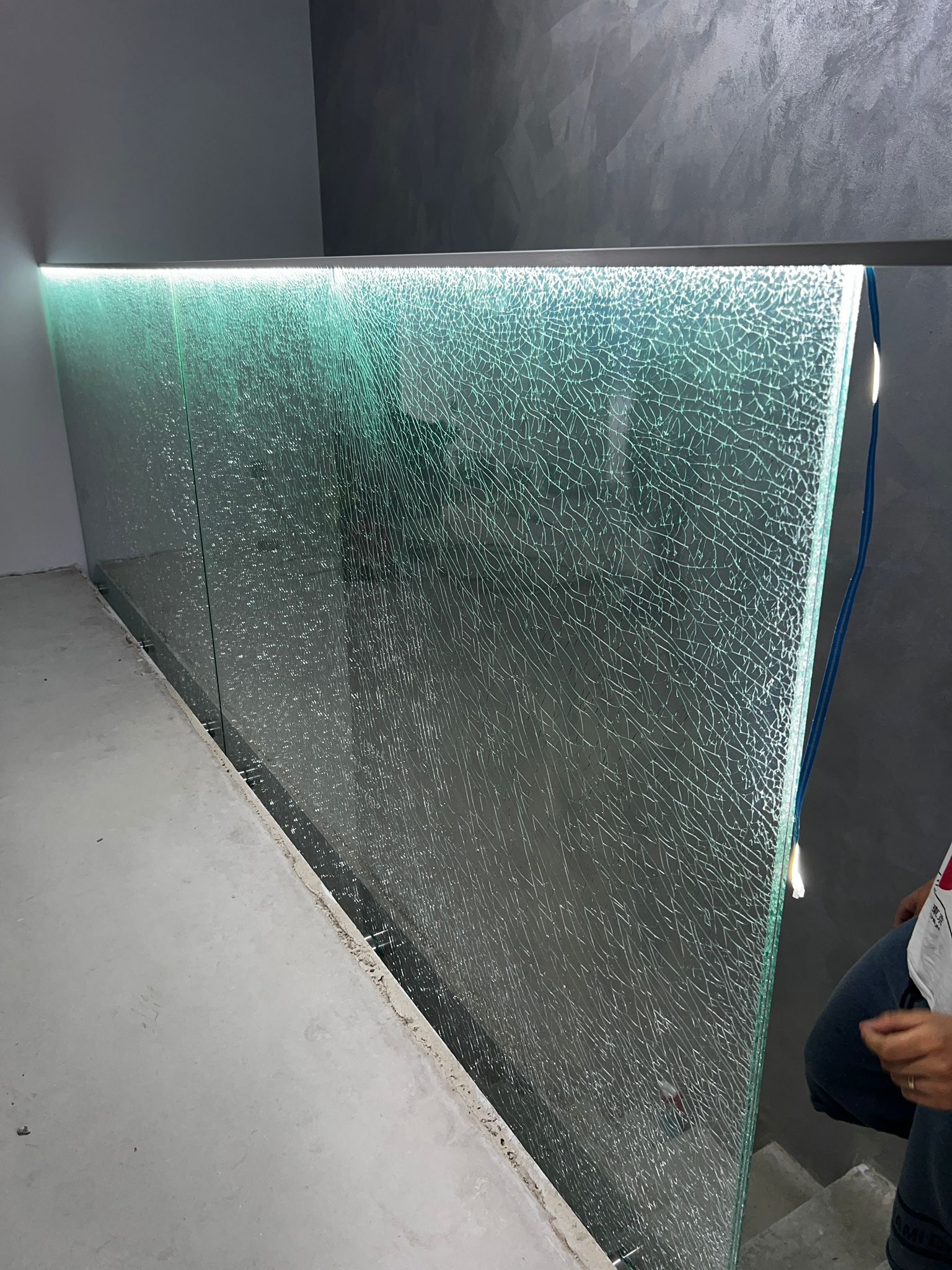 Szklana balustrada z efektem spękania, podświetlona LED, zamontowana przy schodach. Widoczny fragment osoby. Nowoczesny design.