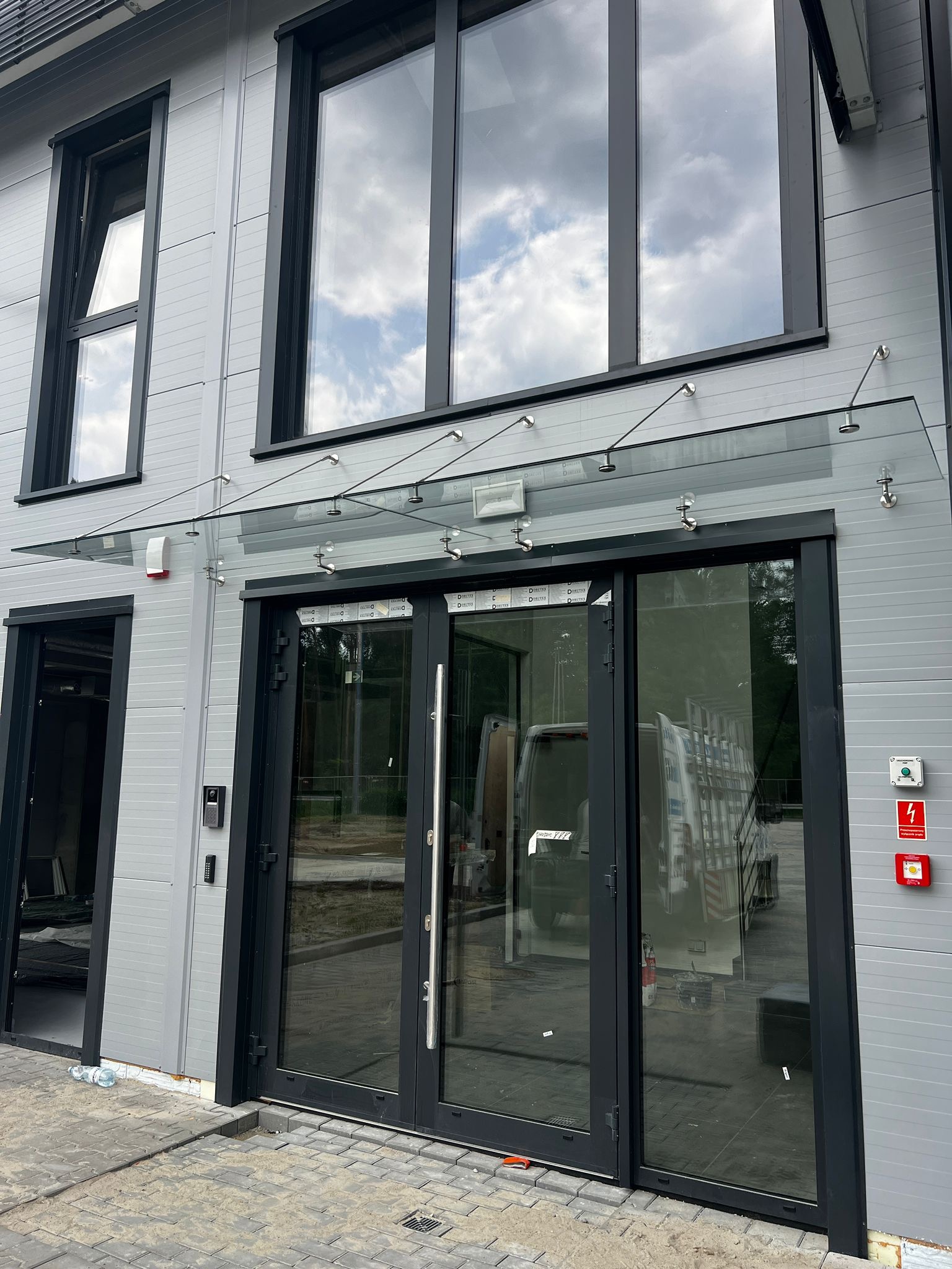 Nowoczesne, szklane zadaszenie nad wejściem do budynku w Kiełczowie. Czarne ramy okien i drzwi kontrastują z szarą elewacją. Widoczne mocowania ze stali nierdzewnej.