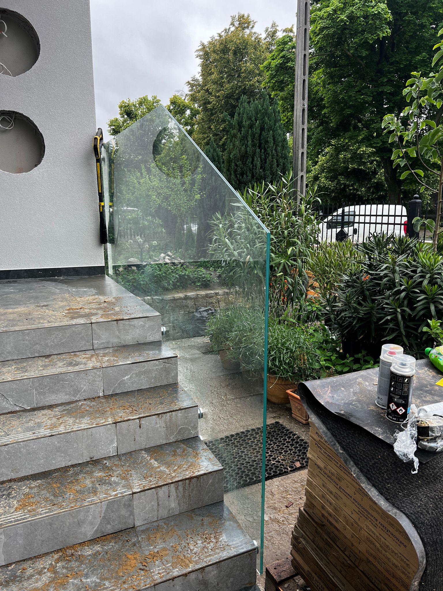 Montaż szklanej balustrady przy schodach wejściowych. Widoczne narzędzia i materiały budowlane. Tło: zieleń ogrodu i fragment budynku.