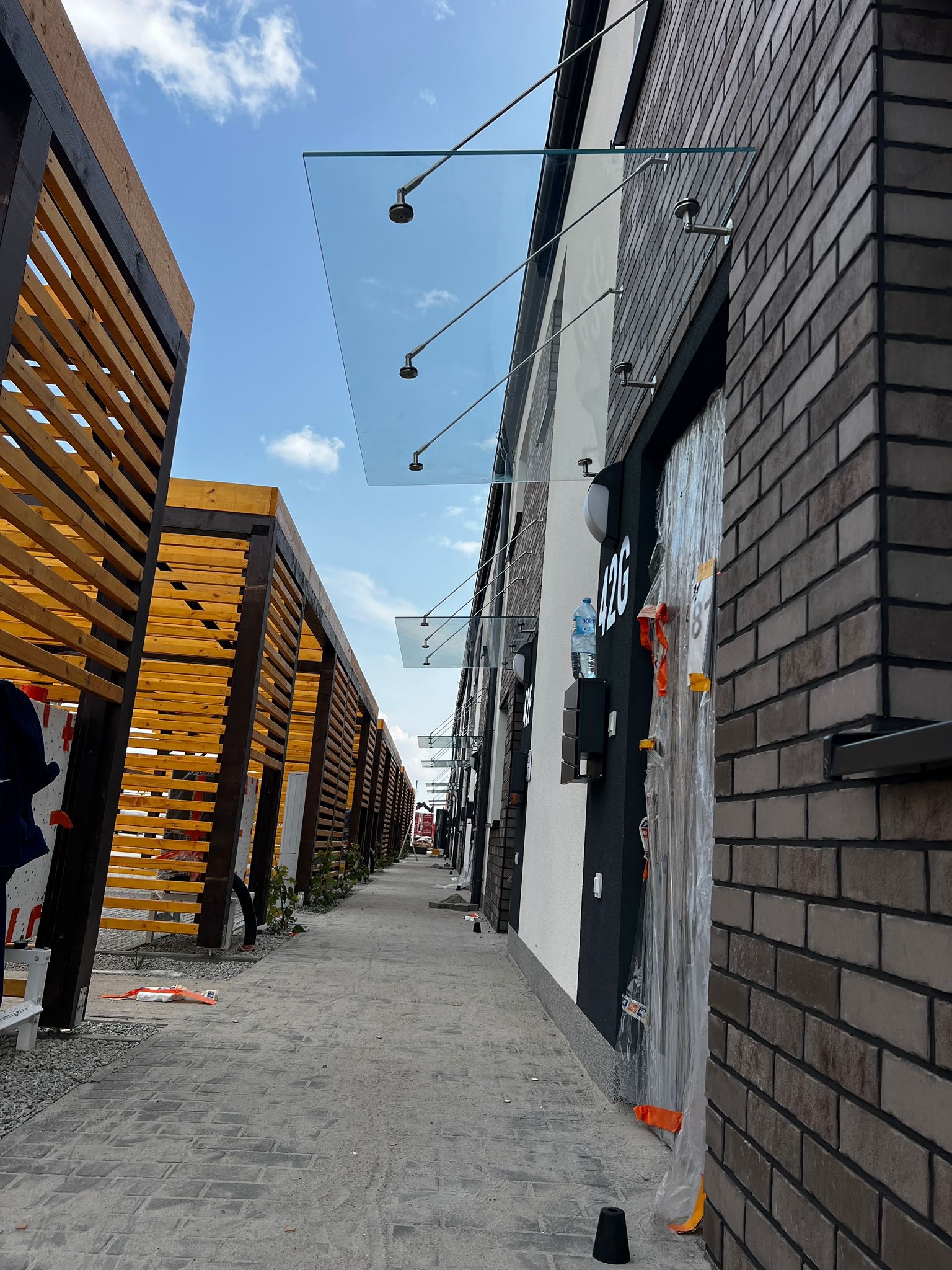 Szklane zadaszenia nad wejściami do budynków z cegły, wsparte na metalowych wspornikach, widoczne z perspektywy chodnika. Po lewej drewniana konstrukcja.