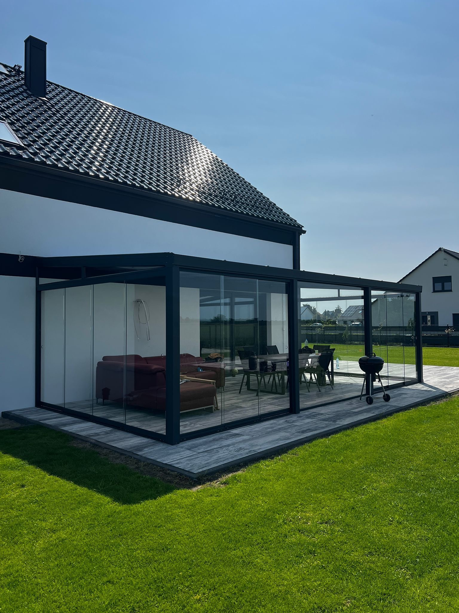 Nowoczesna, przeszklona zabudowa tarasu z czarną ramą, widok z zewnątrz na meble wypoczynkowe i stół. Grill na tarasie, trawnik i dom w tle.