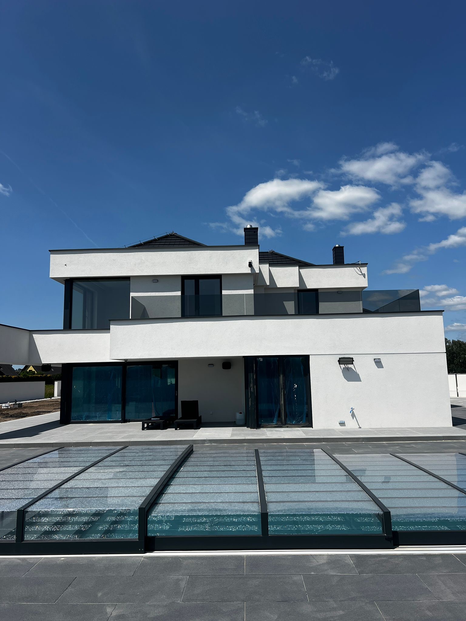 Nowoczesny dom z przeszkloną balustradą na dachu i zadaszeniem basenu z przezroczystego szkła. Błękitne niebo z chmurami w tle. Minimalistyczny design.