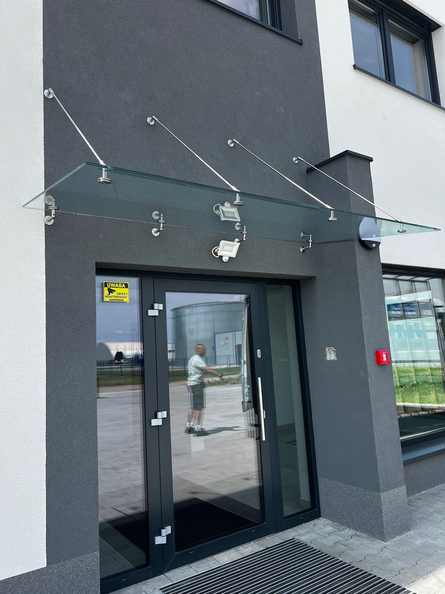 Szklany daszek nad wejściem do budynku, wsparty na metalowych wspornikach. Widoczne drzwi z szybą, tabliczka informacyjna i fragment elewacji w odcieniach szarości i bieli.