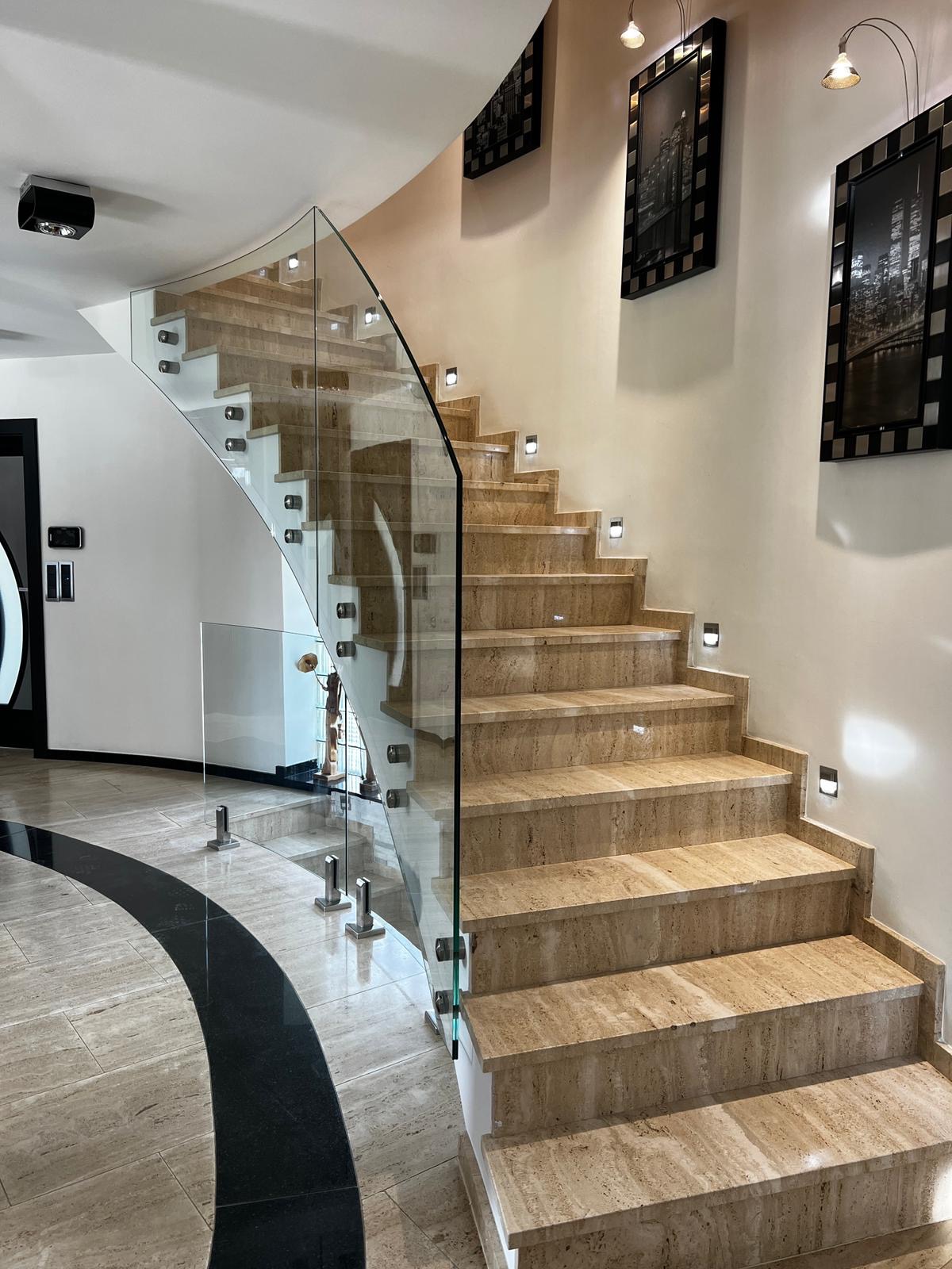 Eleganckie schody z trawertynu z nowoczesną szklaną balustradą, podświetlane stopnie, czarne ramy obrazów na ścianie, widok z dołu.