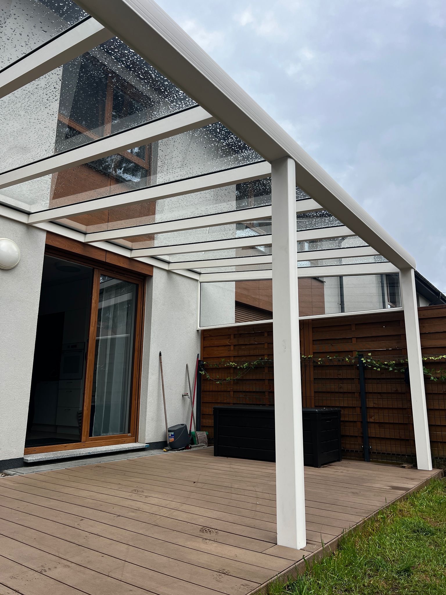 Zadaszenie tarasu z przezroczystego szkła, wsparte na białych, prostokątnych słupach. Widoczne krople deszczu na szklanej powierzchni. Drewniany taras i fragment ogrodu.