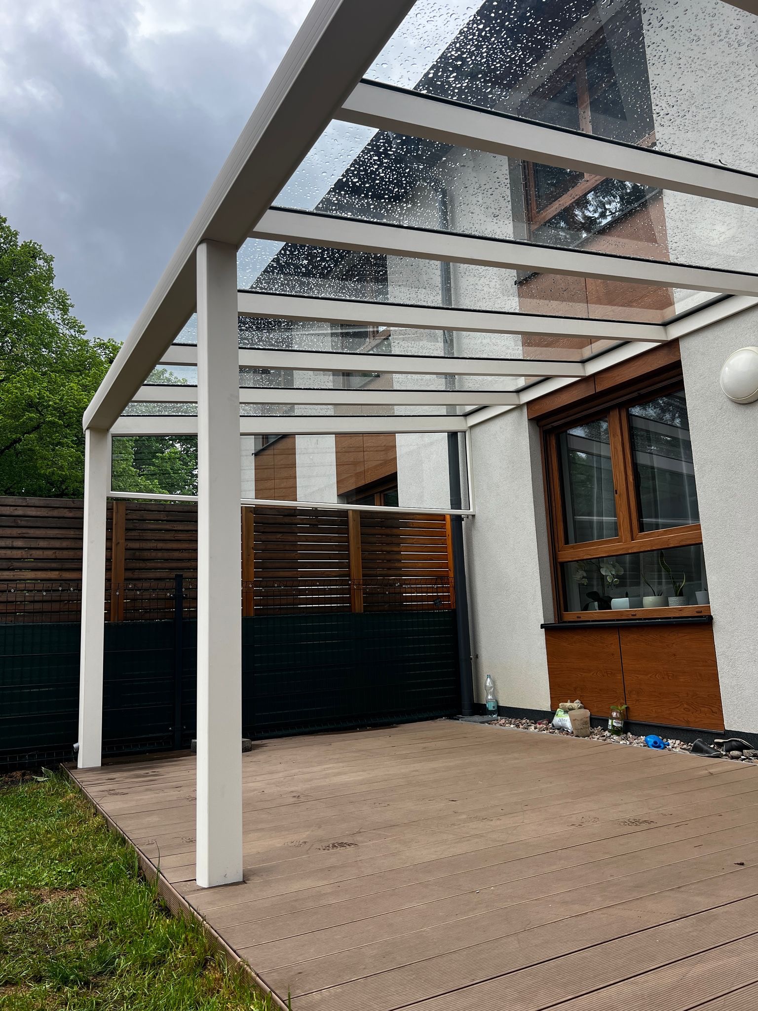 Zadaszenie tarasu z przezroczystego szkła, wsparte na białych, prostokątnych słupach. Widoczne krople deszczu na szklanej powierzchni i drewniana podłoga tarasu.